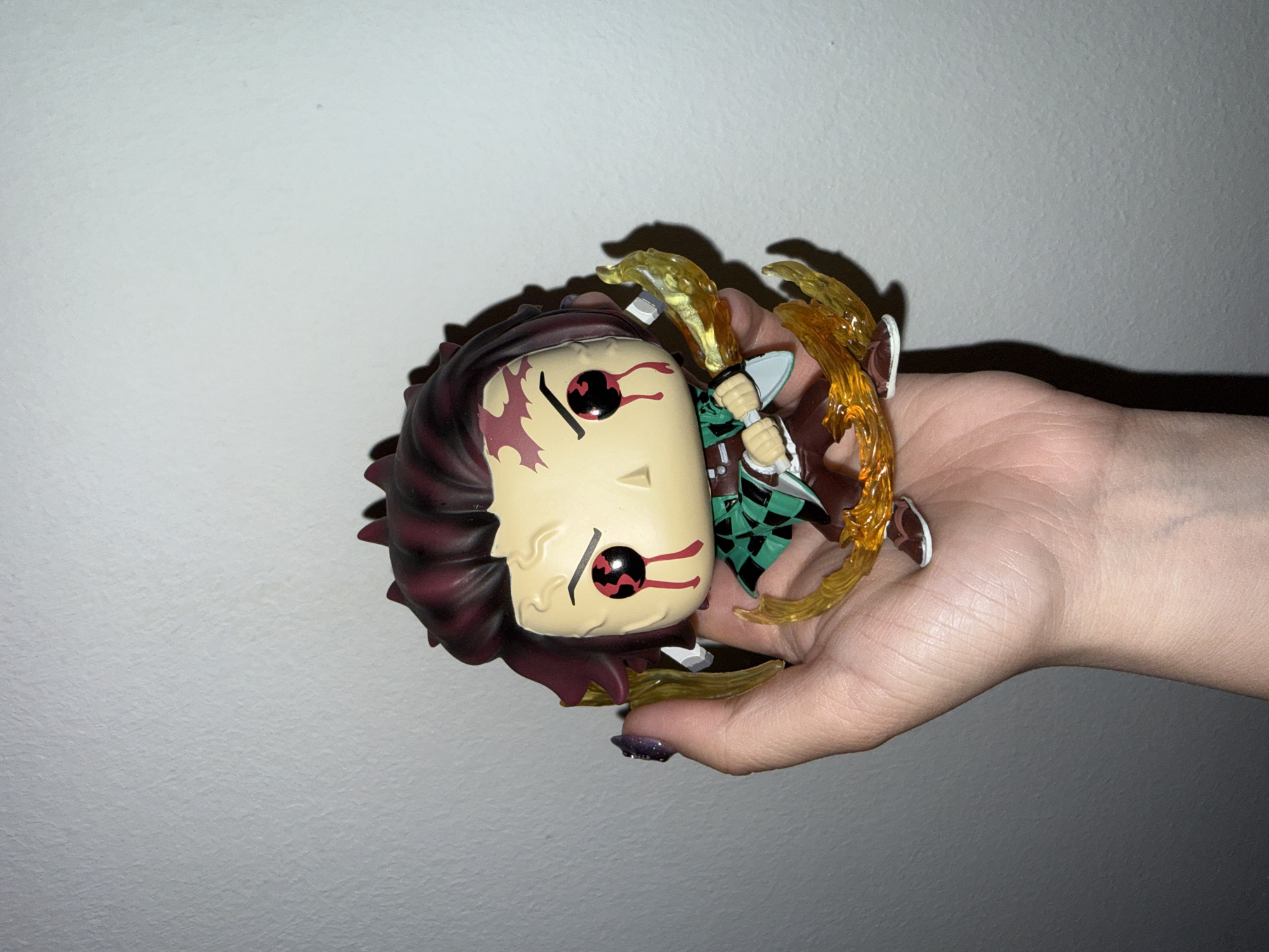 An adult's hand holds a collectible Funko Pop! figure against a plain, light-colored wall in what appears to be an indoor setting in Zagreb, Croatia. The image is oriented sideways, but if viewed upright, the hand is extended horizontally, presenting the figure.
The figure is an action-posed depiction of a character, likely Tanjiro Kamado from the anime *Demon Slayer*, specifically in a demonized or agitated state. It features dark red, spiky hair, a beige face with large, striking red eyes with black pupils, and dark red, dripping markings resembling blood or demon veins under the eyes and on the forehead. The character wears a green and black checkered haori or uniform over a brown base. Translucent yellow and orange flame-like or energy effects emanate from around the figure's arms, suggesting an active attack or power manifestation.
The hand holding the figure is light-skinned, with the thumb showing dark purple, glittery nail polish. Strong directional lighting from an unseen source casts distinct shadows of the hand and the figure onto the textured wall, creating contrast and depth. The wall itself is a uniform, light grey-white color with a subtly rough or stucco texture. No specific location cues for Zagreb are visible within the image frame beyond the general indoor environment. There is no visible text. The time of day is not discernible, but the lighting suggests artificial indoor light.