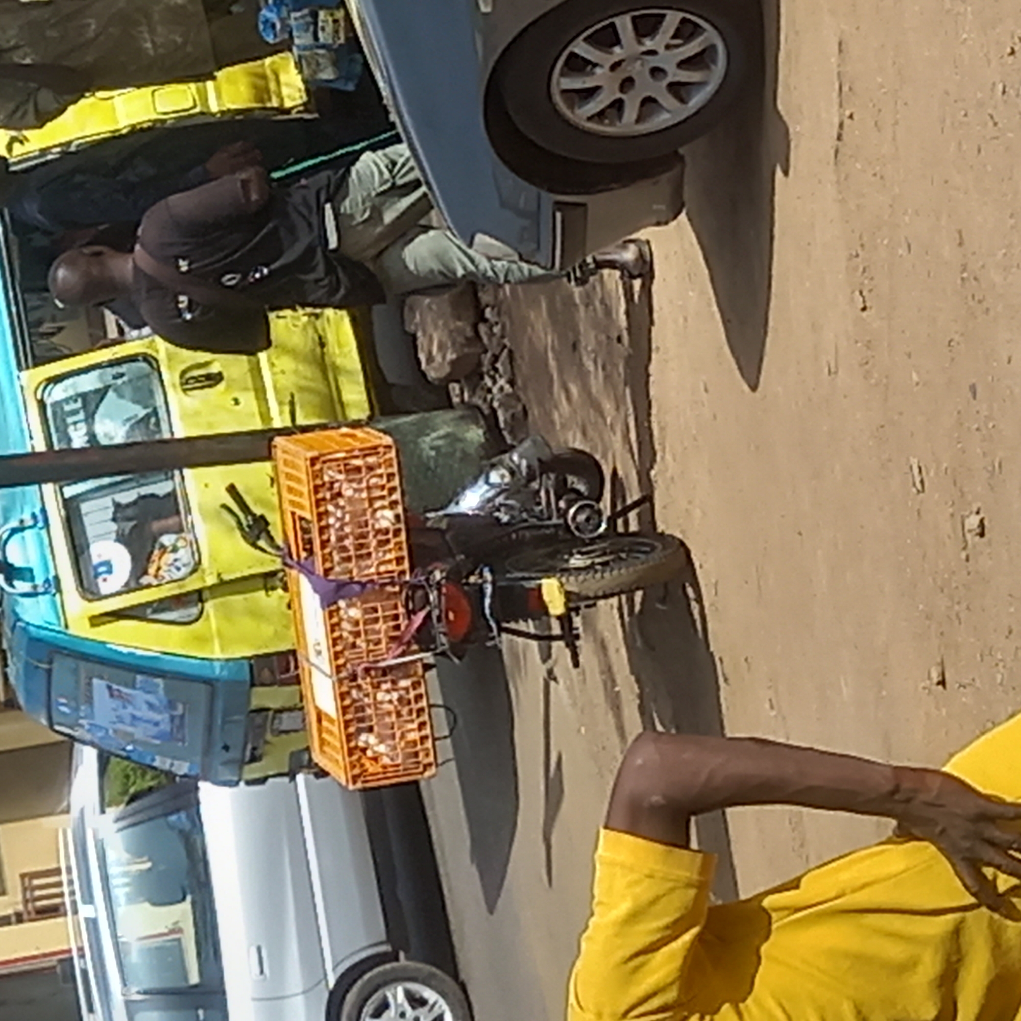 The image depicts a bustling street scene in what appears to be Dadi Riba, Nigeria, characterized by bright daylight and dry conditions. The ground is unpaved, consisting of reddish-brown dirt and small rocks, with long, distinct shadows cast by vehicles, suggesting either morning or late afternoon.
In the mid-ground, a black motorcycle is prominently featured, heavily laden with two orange plastic crates secured to its rear. These crates appear to be filled with numerous small, light-colored objects, possibly eggs or other produce, indicating commercial activity. Adjacent to the motorcycle, a bright yellow and blue minivan or small bus, commonly used for public transport, is parked. Its windows reflect the surroundings, and a person's head is visible inside.
Standing and leaning against the side of the yellow vehicle, near its front, is a man with dark skin, wearing a dark t-shirt and light-colored trousers. He appears to be observing or waiting. Part of a silver/grey car is visible next to him, with its wheel resting on the unpaved ground. Further in the background, another white vehicle is partially discernible.
In the foreground on the right, the arm and torso of another individual wearing a bright yellow t-shirt are visible. Their posture suggests they are either resting or observing the scene.
The overall scene conveys a typical day in a community, with transport, commerce, and daily activities taking place outdoors. No clear, legible text is visible on any of the vehicles or objects in the image.