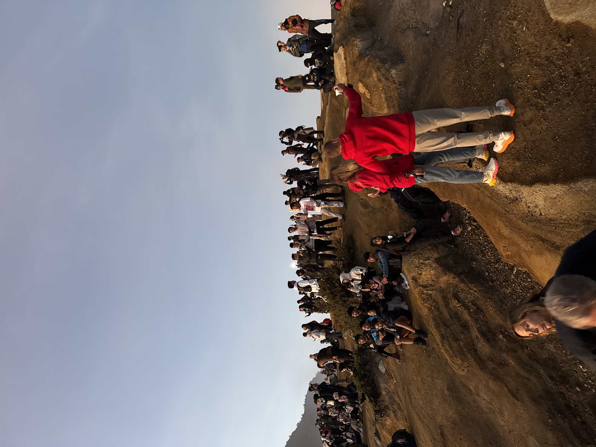 The image shows a crowd of people standing on a hillside in the city of Unknown, Indonesia. The ground is covered in dirt and rocks. The sky is a light blue color. Two people in red shirts stand at the edge. Many people are looking towards the same direction.