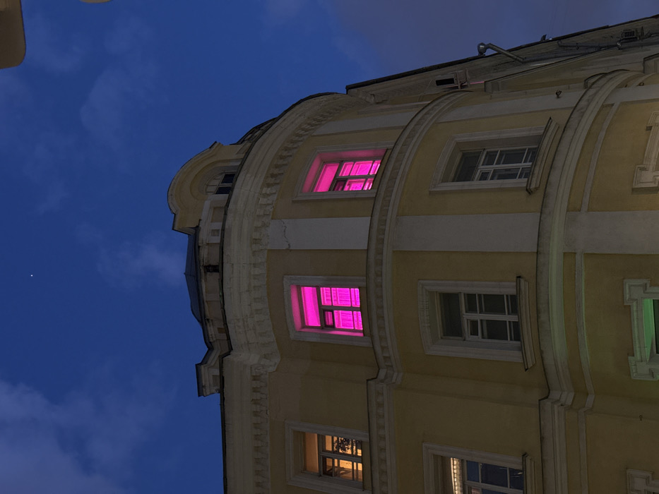The image depicts the exterior of a multi-story building at dusk or night, located at Malaya Bronnaya Ulitsa, 20с1, Moscow, Russia. The building features a light yellow-cream facade with classical architectural elements, including prominent cornices and moldings, and a distinctive rounded corner. A darker-colored roof and an arched attic window are visible at the apex. Two windows on the second floor (from the top) of the rounded section are illuminated with an intense, uniform magenta light. An adjacent window to the left displays a warm orange-yellow light, revealing interior foliage. Lower-level windows are unlit or dimly lit. A dark, vertical drainpipe is visible on the building's right-hand facade. The sky is a deep twilight blue with faint, wispy clouds, and a single bright celestial object (star or planet) is discernible in the upper-left quadrant. No individuals or discernible actions are present in the frame.