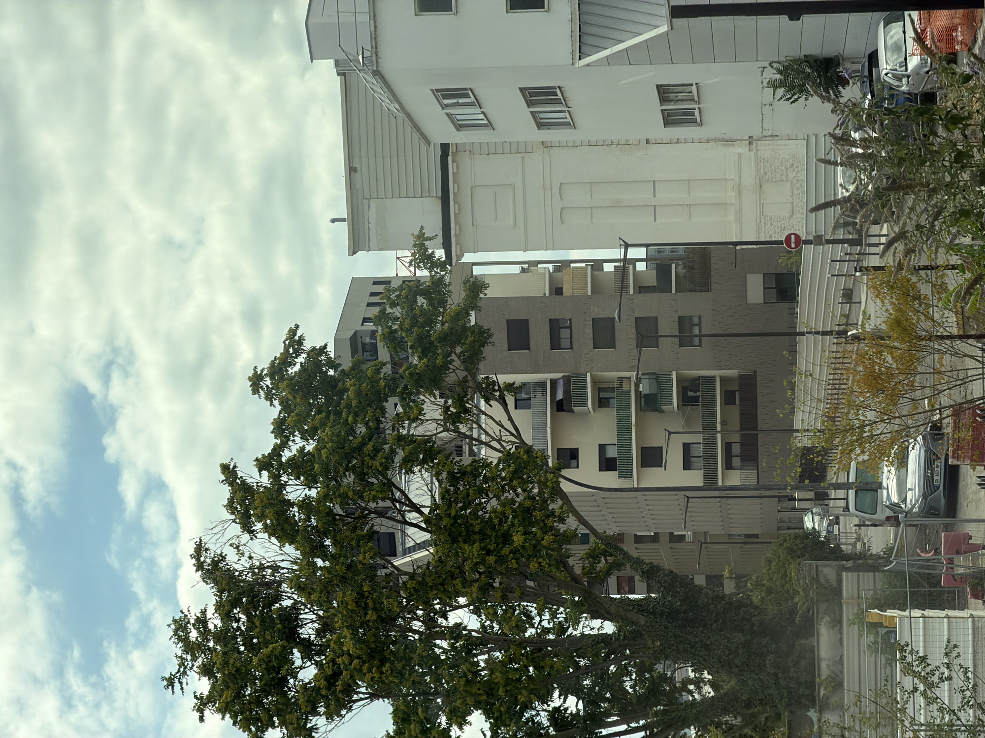 This media file depicts a street scene in Ivry-sur-Seine, France, characterized by modern architecture and greenery. In the foreground, a large tree with dense green foliage dominates the left side, its branches extending upwards. Behind the tree, multiple buildings are visible, showcasing a mix of architectural styles.
The buildings appear to be residential or mixed-use, with light-colored facades and numerous windows. One building in the background features a grey, textured facade with darker rectangular windows, and balconies with horizontal railings. Further back, a white building with a more traditional design is present, with rows of windows. The architecture suggests a contemporary urban environment.
Several vehicles are parked along the street. A silver SUV with a license plate is prominently featured on the right side. Other vehicles, including a white van, are also visible in the background. The presence of streetlights and the general layout suggest a public street or residential complex.
A red circular sign with a white horizontal bar is mounted on a pole, indicating a "no entry" or similar traffic restriction. Construction or renovation elements, like fencing and what appear to be temporary structures, are also present, suggesting ongoing development or maintenance in the area.
The sky is overcast with scattered clouds, indicating a potentially cool or temperate day. The lighting suggests it could be daytime, possibly late morning or afternoon, with diffused light due to the cloud cover. The overall mood is neutral, capturing a snapshot of urban life with a blend of built environment and natural elements. No people are directly visible in the frame.