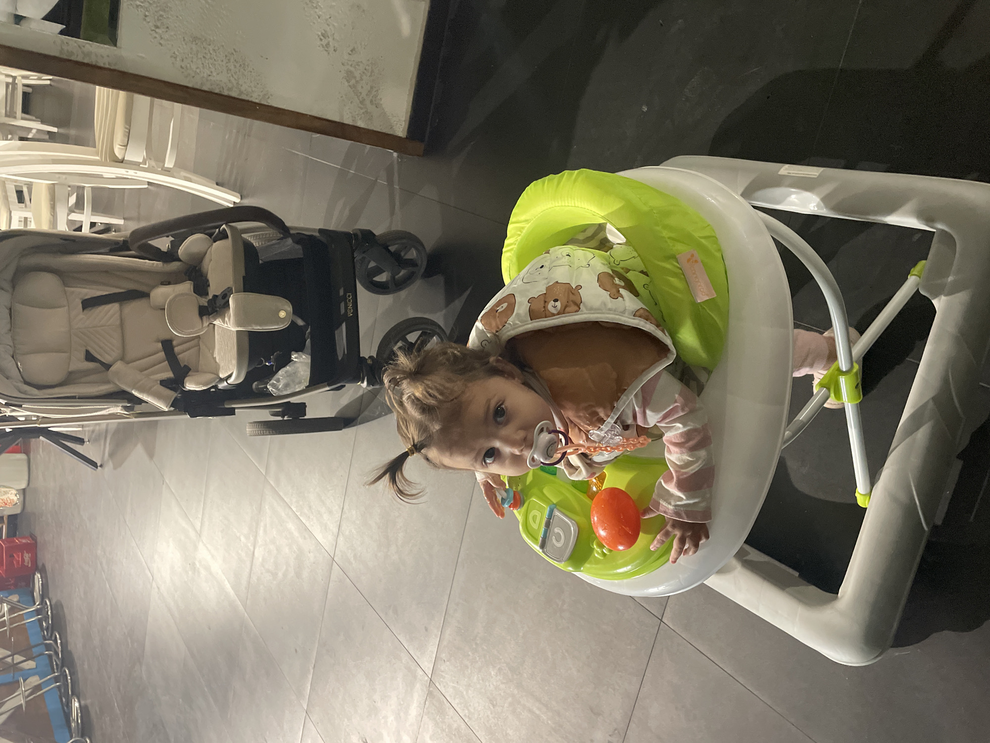 An overhead shot captures a young child in a baby walker indoors, with a stroller visible in the background. The child, likely a toddler, has light brown hair styled in two small pigtails and is looking upwards directly at the camera with a neutral, curious expression. A pacifier is in their mouth. They are wearing a light-colored hooded top adorned with brown bear patterns, over a striped long-sleeved shirt.
The baby walker is primarily white and bright lime green, featuring an orange toy on its front tray, which the child's right hand is near. A small pink tag with partially visible text, possibly "Kangaroo," is attached to the green fabric of the walker. The stroller in the background is beige or light cream with a black frame and wheels. The brand "VENICCI" is distinctly visible on its side frame.
The setting appears to be an indoor space with large, light grey tiled flooring that reflects some light. In the background, there are numerous white chairs, some stacked, suggesting a dining area or a space where furniture is being stored. The lighting is artificial and creates some shadows, implying an evening or nighttime scene.