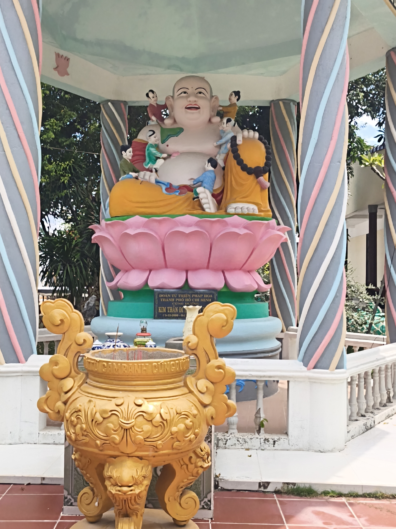 The image depicts an outdoor scene at a Buddhist temple in Cam Ranh, Vietnam, as indicated by the inscription "CAMRANH GUNG DUONG" on the golden incense burner. In the foreground, a large, ornate golden incense burner with intricate carvings and clawed feet sits on a tiled floor. Behind the incense burner, a colorful statue of a laughing Buddha is prominently displayed, seated on a pink lotus blossom. The Buddha statue is adorned with small figures of children. The statue is housed within a pavilion supported by pillars painted with diagonal stripes of various colors, including pink, yellow, blue, and gray. A white railing runs along the front of the pavilion. Trees and foliage are visible in the background, suggesting a tropical environment. The overall scene is brightly lit, indicating daylight.