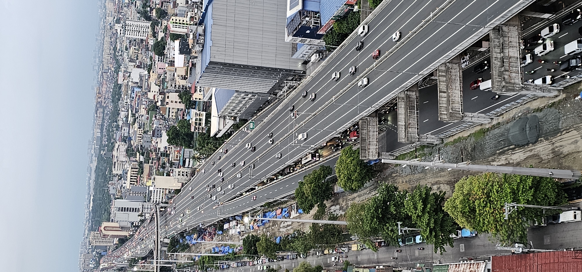 This is an aerial, rotated view of a busy multi-lane overpass in Makati, Philippines, likely during daylight hours. The overpass carries a significant amount of traffic, with numerous cars and some trucks visible on its lanes. The sky is hazy and overcast, suggesting a cloudy or polluted atmosphere.
Beneath the overpass, the scene is a stark contrast. A densely populated urban area with numerous buildings of varying sizes and architectural styles stretches into the distance. Interspersed with the buildings are pockets of green trees. At ground level, directly adjacent to the overpass supports, there appears to be a makeshift settlement or informal housing area, evidenced by numerous tents or tarpaulin structures in blue and other colors. Power lines and poles are visible throughout the scene, indicating the presence of infrastructure.
The perspective is high, suggesting the photograph was taken from a tall building. The activity on the overpass is vehicular traffic, while the area below seems to be a mix of residential and informal living spaces. The overall impression is one of urban density, with significant traffic congestion and a juxtaposition of modern infrastructure with informal settlements at ground level. No discernible text is visible in the image.