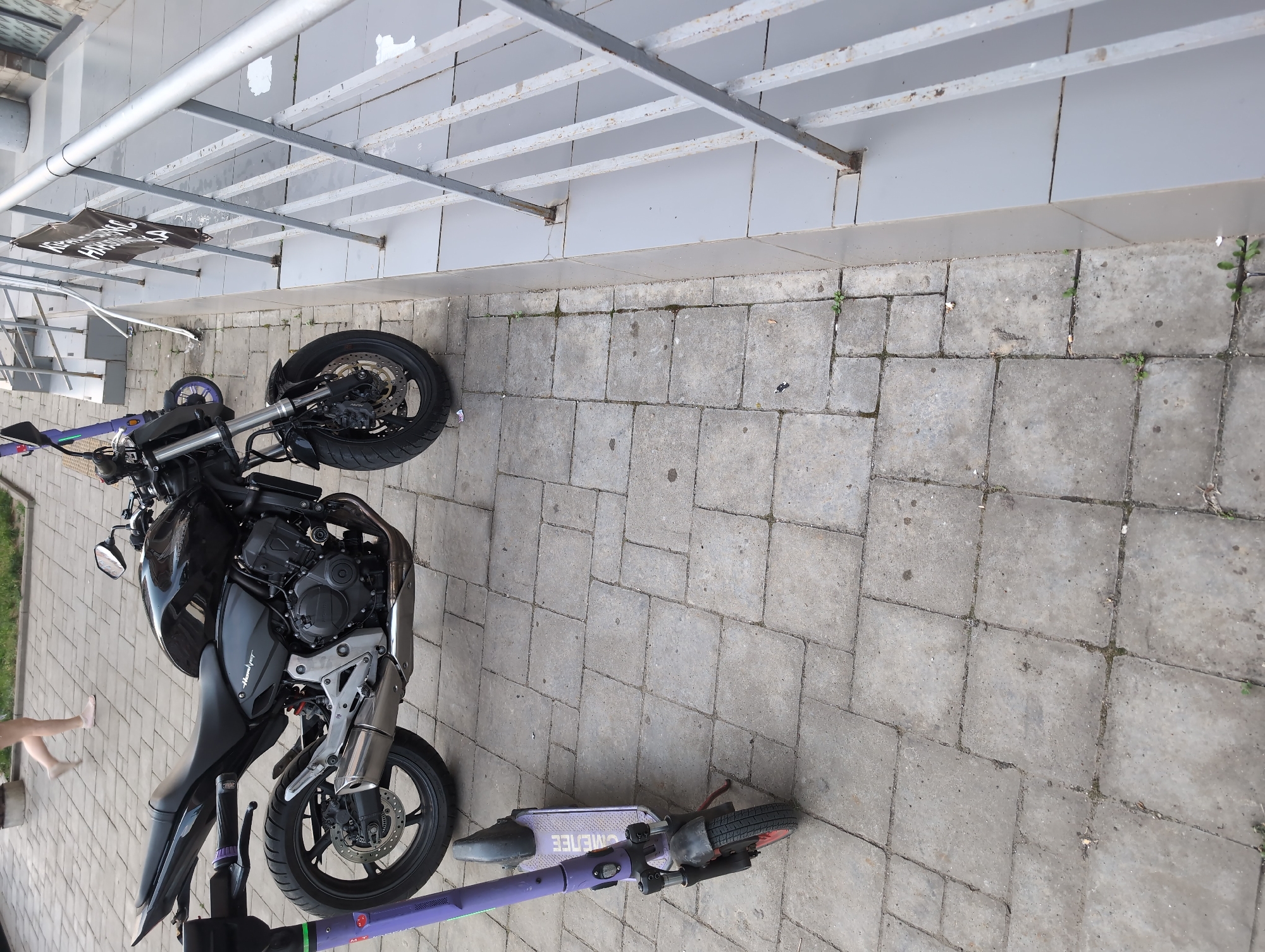 A black motorcycle and a purple electric scooter are parked side-by-side on a paved area with square stone tiles. The motorcycle is positioned to the left, with its front wheel slightly turned. The scooter is to the right, with its handlebars extended. A dark banner with white text is hanging from an overhead metal structure above the motorcycle. Part of a person's legs can be seen on the left side of the image, near the motorcycle. The scene appears to be outdoors, possibly in a public area or parking lot, with a building or wall in the background. The lighting suggests it is daytime. The text on the banner is partially obscured but appears to say "HORSES".