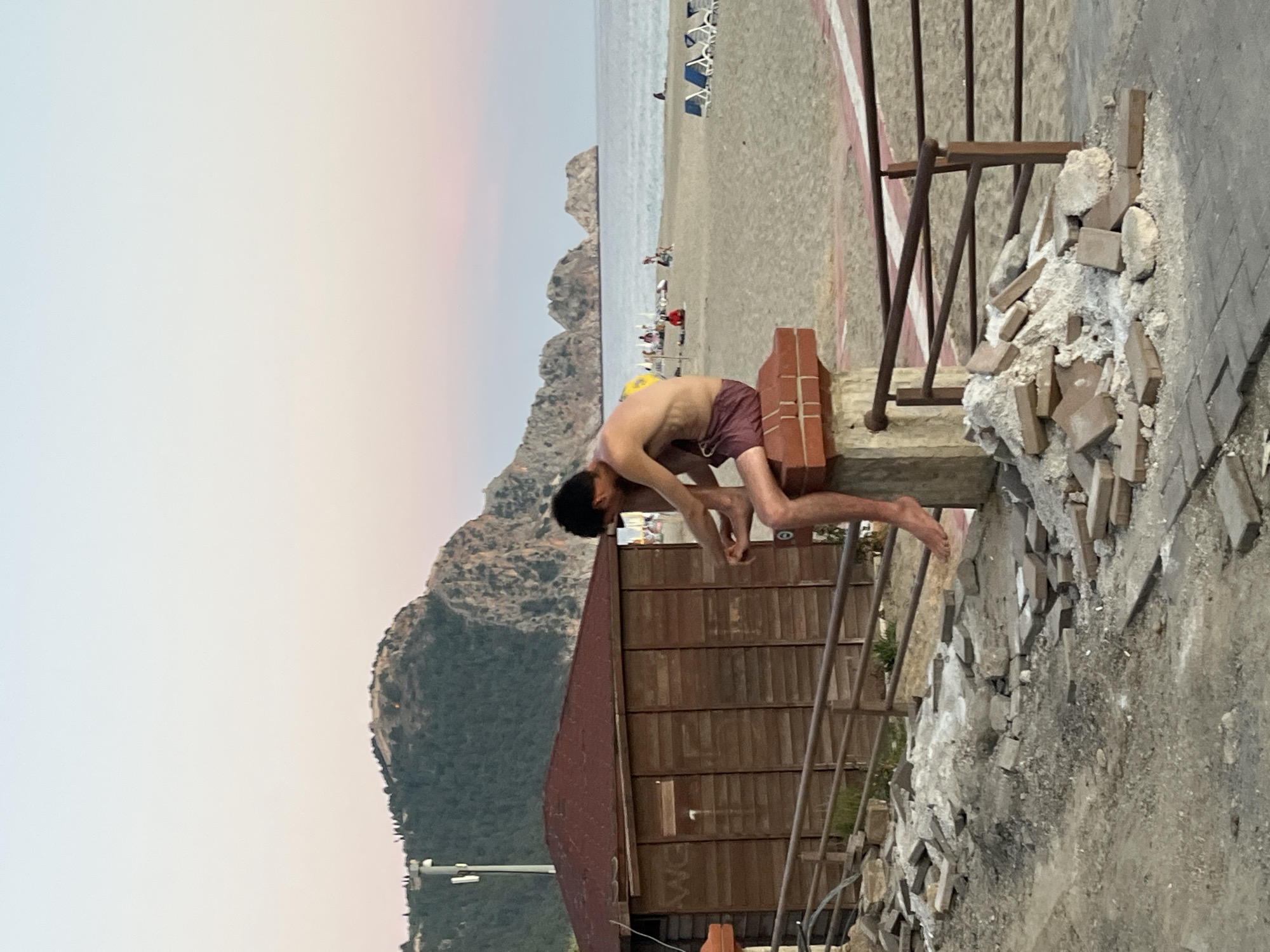 A photograph depicts a man in maroon swim trunks standing on a brick wall adjacent to a building with a red tile roof, overlooking a beach scene. The beach has lounge chairs and people. Mountains are visible in the background, under a sky transitioning from light to dark. The man is bending down, possibly washing his hands or feet. Rubble, consisting of bricks and concrete, is scattered around the structure. A metal railing runs alongside the wall. The photograph was taken in the city of Alanya, Turkey.