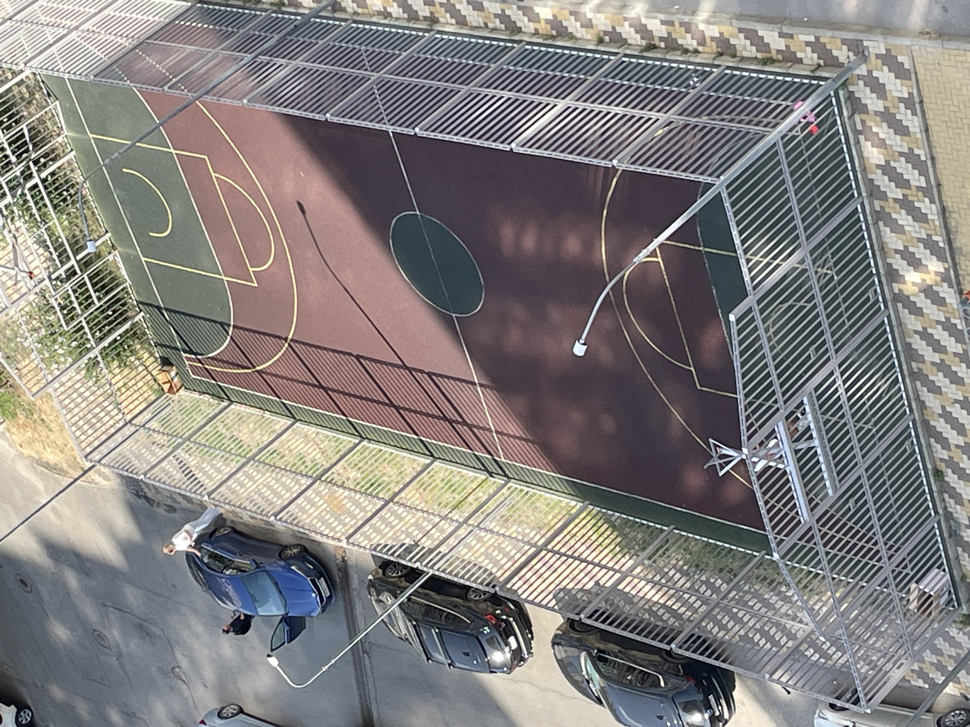An overhead shot captures a basketball court fenced with metal bars. Two cars, a blue one with its door open and a black one behind it, are parked near the court. A person in a white top and light-colored pants stands next to the blue car. The court has a dark brown surface with green sections and yellow markings for basketball court lines. A street lamp is visible near the court. The surrounding area is paved with patterned tiles.