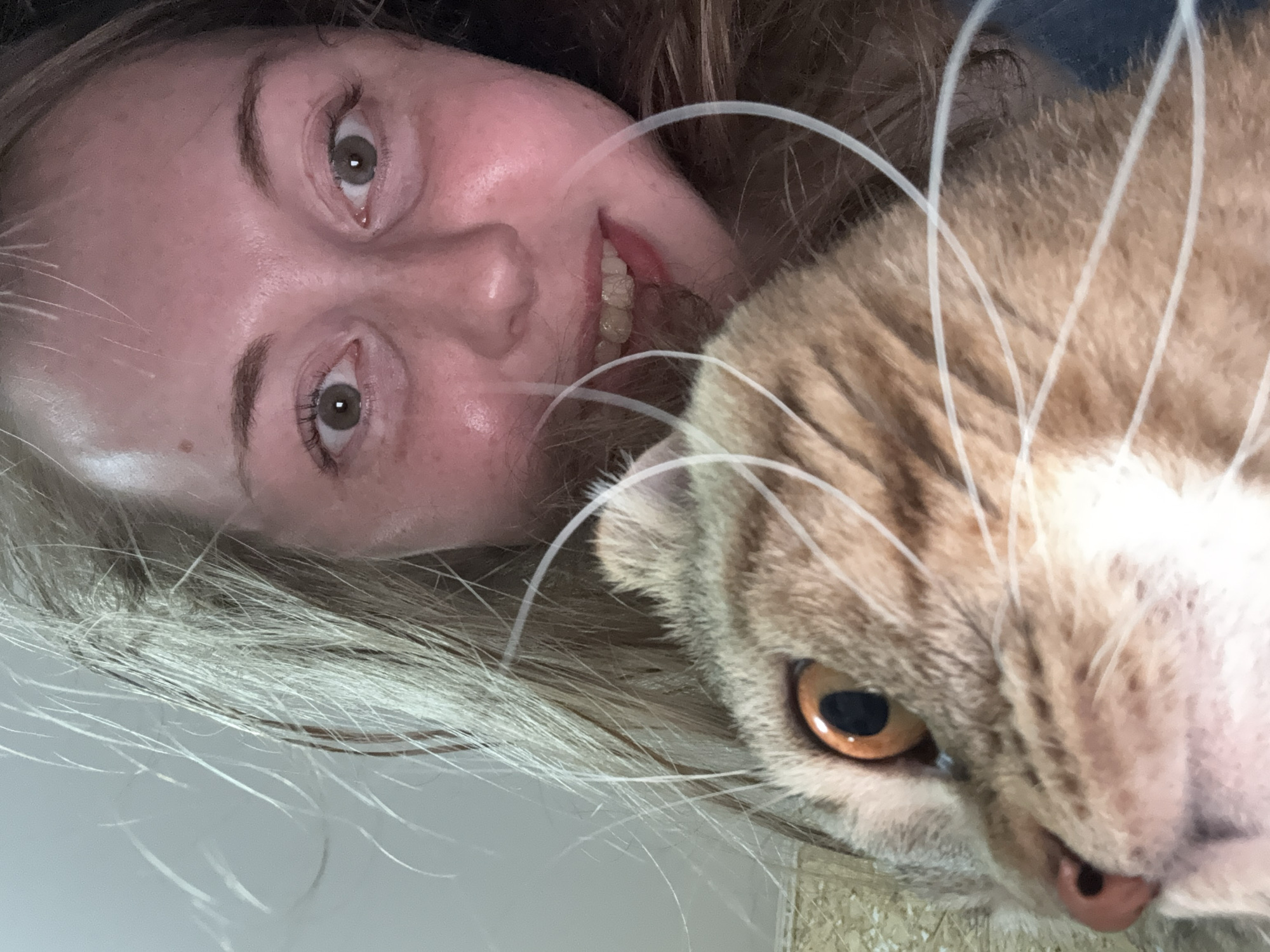 A close-up selfie shows a young woman and a cat. The woman, with long blonde hair and fair skin, is looking at the camera with wide green eyes and a slight smile. Her face is slightly flushed, and she has freckles. The cat, a tabby with orange and brown fur, is positioned in the foreground, partially obscuring the woman. Its large, orange eye is in sharp focus. The background is nondescript, with a suggestion of a wall or furniture. The image appears to have been taken indoors.
