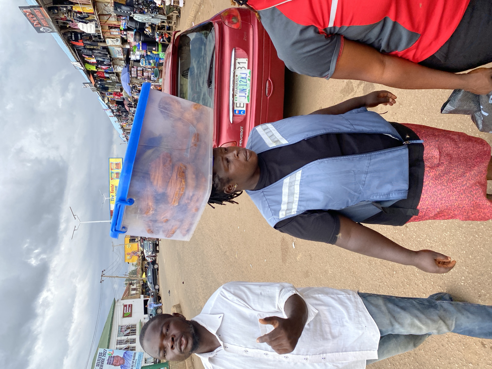 The image shows a street scene in Bukuru, Nigeria, with two men and a red Peugeot car. The man in the foreground is wearing a white shirt and blue jeans, looking towards the camera with a slight smile and making a hand gesture. He appears to be outdoors, possibly in a market or busy street, given the background which shows stalls with goods and other vehicles.
Behind him, another person, who appears to be a woman, is carrying a clear plastic container filled with food items on her head. She is wearing a blue vest over a black shirt and a patterned red skirt. Her expression is neutral. The red Peugeot car, with a Nigerian license plate visible (E JJN-12AJ 31, Plateau), is parked nearby.
The background reveals a bustling environment with shops, stalls displaying various items like clothing and food, and other vehicles. Billboards with text are also visible, including one that reads "SELFLESS SERVICE 2027" and another mentioning "The Taste HOME." The sky is cloudy, suggesting it might be an overcast day. The overall impression is a candid shot capturing everyday life in a busy Nigerian setting.