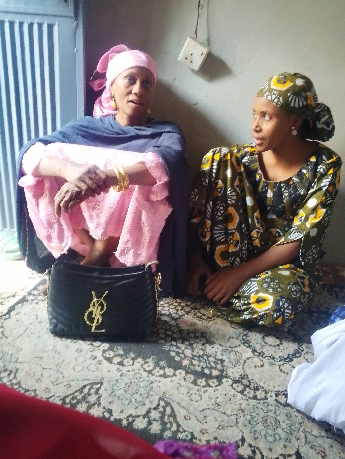 Two adult females are seated on a patterned rug in an indoor setting in Dorayi, Nigeria. The woman on the left, appearing older, wears a pink headscarf, a pink garment, and a dark blue shawl. Her bare feet are partially visible, and she has multiple gold-colored bangles on her right wrist and a gold earring on her visible left ear. Her mouth is slightly open, indicating she is speaking. The woman on the right, appearing younger, wears a patterned olive-green, yellow, black, and white headscarf and matching dress, with a pearl-like earring on her visible left ear. She is looking directly at the woman on the left, with her hands resting in her lap. Between them, on the rug, rests a black quilted handbag featuring a gold 'YSL' logo and a chain strap. The background shows a plain, light-colored wall with a white electrical outlet and exposed wiring mounted on it. To the far left, a vertical slatted structure is partially visible. A pair of light green sandals is located near the feet of the woman on the left. White fabric is present on the rug to the far right, and colored fabric is in the immediate foreground.