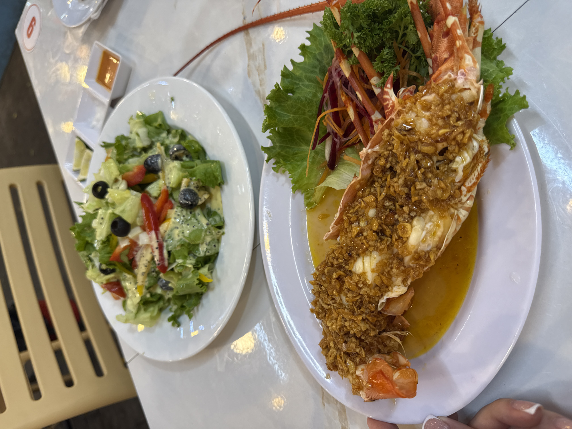 The image displays an overhead view of a dining table set with two plates of food and condiments, captured in a restaurant setting in Phú Quốc, Vietnam.
The dominant feature is a large, halved lobster served on a white oval plate, positioned on the right side of the frame. The lobster meat is generously covered with a layer of golden-brown, crispy fried garlic or shallots, and it rests in a pool of yellowish sauce. Fresh green lettuce, curly parsley, and thin strips of red and purple vegetables garnish the plate around the lobster. The spiny antenna of the lobster extends into the upper left background.
To the left, a round white plate holds a vibrant green salad. This salad features fresh lettuce, black olives, and slices of red and yellow bell peppers, all coated in a creamy, light-colored dressing containing small dark specks.
In the upper left corner of the table, small white rectangular dishes are visible. One contains an orange-brown dipping sauce, while another appears to hold lime wedges. A person's hand, adorned with a white-tipped manicure, is visible in the bottom right, gently holding the edge of the lobster plate.
The table surface is a glossy, light-colored material, showing reflections of ambient lighting. Part of a light-colored, slatted chair is partially visible on the far left. A red number "6" within a white circle is visible on the table surface in the upper left quadrant. The scene suggests a meal, possibly dinner, in an indoor establishment.