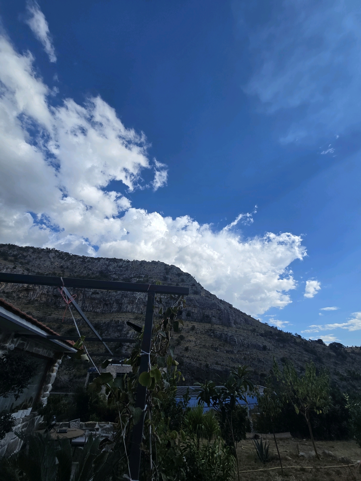 This image depicts a mountainous landscape under a bright blue sky with scattered white clouds. In the foreground, there is a portion of a building with a tiled roof, a stone wall, and an outdoor seating area with a table and chairs partially obscured by foliage. A metal structure with a wire mesh roof is also visible, with vines growing on it. Beyond this, a lush green area with various trees and plants leads towards a blue-tiled swimming pool. The background is dominated by a rugged, rocky mountain slope covered with sparse vegetation and trees. The scene appears to be a residential area or a rural retreat situated at the base of a mountain. While the image captures a scenic natural environment, there are no people visible. The location is identified as being in Podgorica, Montenegro.