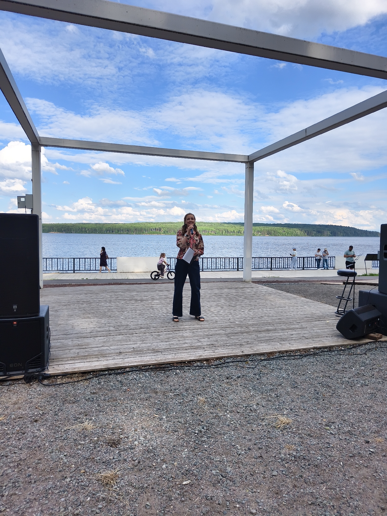 A woman is speaking into a microphone on a wooden stage. She is wearing a patterned shirt and dark pants. The stage is outdoors, with a large body of water and a forested shoreline in the background. The sky is blue with white clouds. To the left of the stage, a large black speaker is visible. To the right, a stool is placed in front of another speaker. In the background, several other people are present, including a woman walking, a child on a bicycle, and a group of people standing and talking. The scene appears to be a casual outdoor event. The time of day appears to be daytime, with bright sunlight. The weather is pleasant, with a blue sky and some clouds. There is text visible on the speaker on the left that reads "JBL" and "EON".