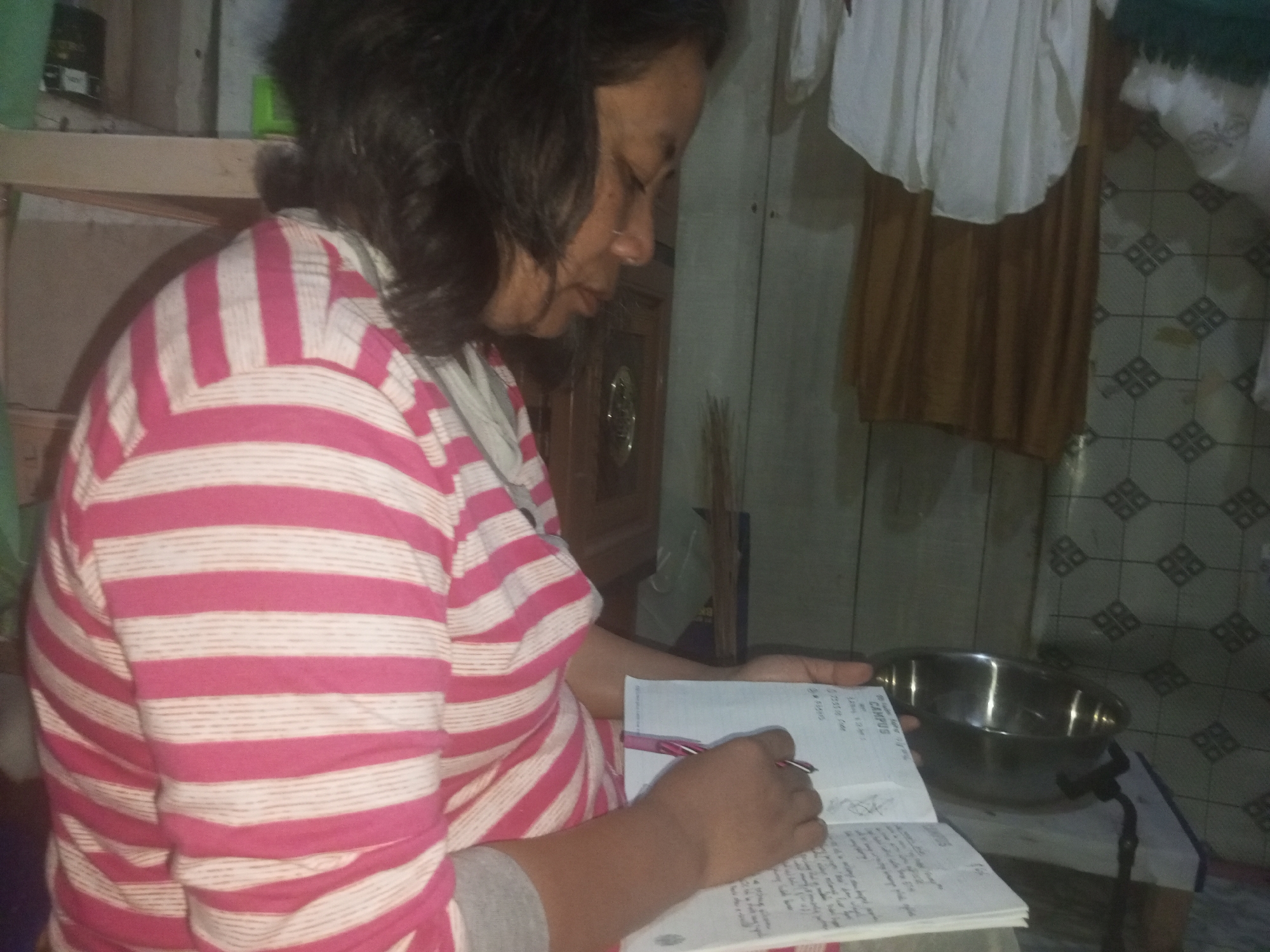 In a dimly lit, cluttered room, a person is seated and focused on a notebook, writing with a pink pen. The person is wearing a pink and white striped long-sleeved shirt and a grey undershirt. A metal bowl is placed on a surface next to the notebook. The background reveals a tiled wall and a wooden cabinet. The setting appears to be a residential interior in Andongsili, Indonesia, likely a humble dwelling given the visible disarray.