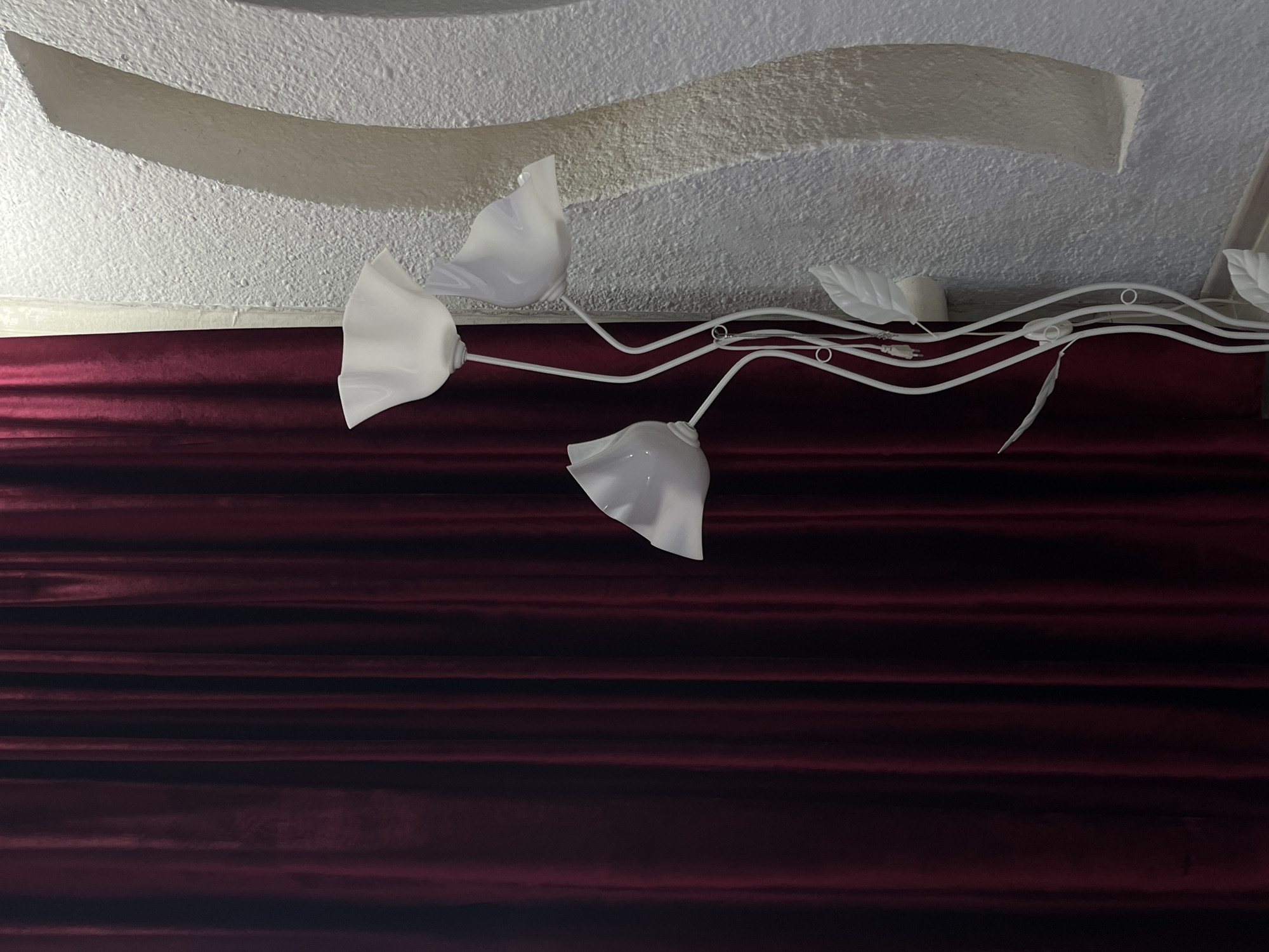 A floor lamp with three white, flower-shaped lights stands to the right of a dark red curtain. The lamp has a white, vine-like stem with several leaf-shaped accents. The wall behind the lamp is textured and white, with a curved architectural detail. The scene appears to be indoors, possibly in a residential or commercial space.