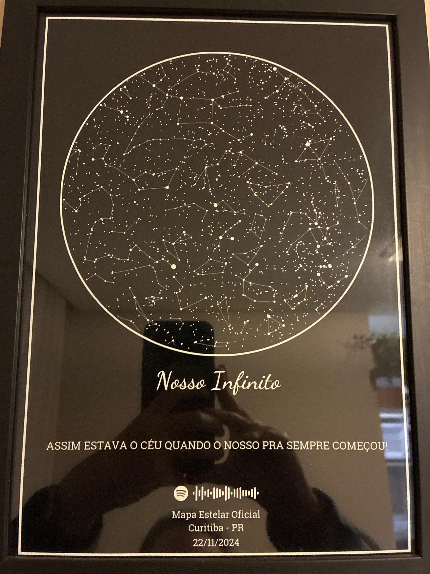 The image shows a framed star map with constellations displayed within a circle on a black background. Below the star map, text reads "Nosso Infinito" in a stylized font, followed by "ASSIM ESTAVA O CÉU QUANDO O NOSSO PRA SEMPRE COMEÇOU!". A Spotify logo and a waveform are also visible, with "Mapa Estelar Oficial Curitiba - PR" and "22/11/2024" printed below. The frame is black with a white matting. A reflection in the glass shows a person holding a phone, obscuring a portion of the text and the lower part of the star map. The background environment is dimly lit, suggesting an indoor setting.