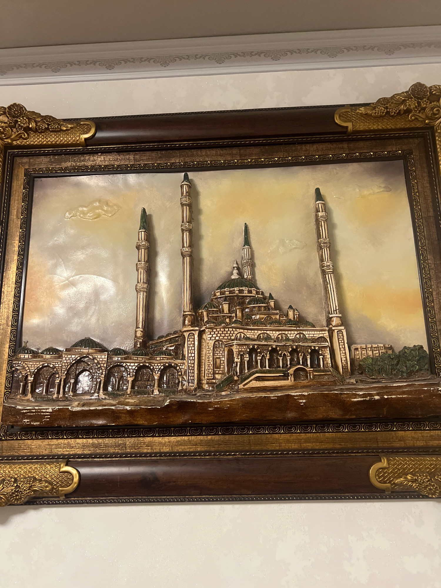 The image displays a framed 3D artwork of a mosque, likely the Grand Mosque in Novyy Kostek, Russia, based on the stylistic similarities of the minarets and domes. The artwork depicts a detailed architectural representation of the mosque with multiple minarets, domes, and an arched colonnade. The mosque is rendered in beige and brown tones with green accents on the domes. The background is a gradient of yellow and grey, suggesting a sky with clouds. The artwork is encased in a multi-layered frame, featuring a dark brown inner frame and a wider, ornate gold outer frame with decorative floral motifs. The artwork is hung on a wall with a textured, light-colored surface and a decorative border at the top. There are no people or animals present in the artwork or the immediate surroundings.