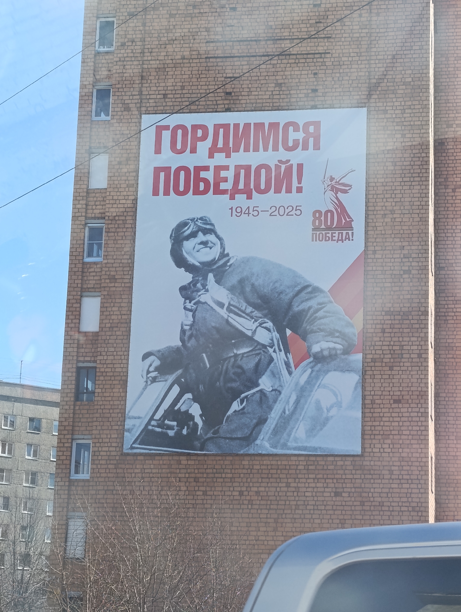The image shows a large commemorative poster attached to the brick facade of a multi-story building. The poster commemorates the 80th anniversary of Victory Day ("Pobeda" in Russian), spanning the years 1945-2025. It features a black-and-white image of a smiling pilot in flight gear within a fighter plane cockpit. The Russian text on the poster reads "Gordimsya Pobedoiy!" which translates to "We are proud of the Victory!". A stylized graphic with a woman holding a sword is also visible.
The building to which the poster is affixed has multiple windows, suggesting it is a residential or office building. Another building is visible in the background on the left side of the image. The foreground includes the top of a silver-colored vehicle, implying that the photo was taken from a car or nearby location. Bare trees are visible in the lower section, possibly indicating late autumn or early spring. Based on the text on the poster, this image is likely taken in the city of Krasnoyarsk, Russia, as Victory Day commemorations are common in Russian cities.