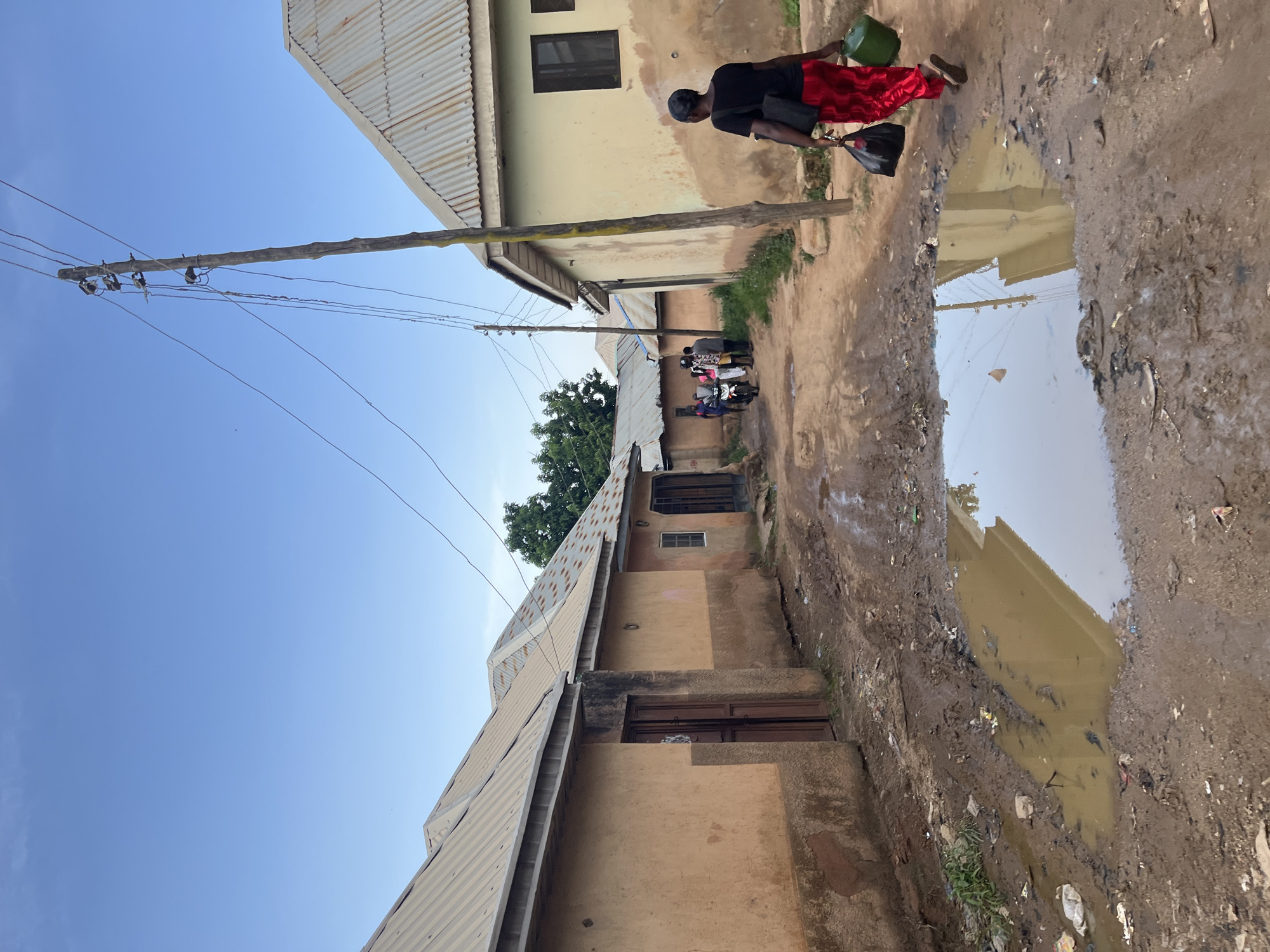 The image depicts a scene in Rafin Par, Nigeria, under a clear blue sky, suggesting it is daytime. The setting is a muddy, unpaved street lined with buildings, some with corrugated metal roofs and light-colored walls. A large puddle of murky water reflects the sky and a building.
In the foreground, a person wearing a black shirt and a bright red skirt is walking away from the camera, carrying a green bucket in one hand and a black bag in the other. Their posture suggests they are engaged in a daily task, possibly collecting water or transporting goods. Further down the street, a group of people are gathered around several motorcycles, some of whom appear to be wearing brightly colored clothing. This suggests a community gathering or people preparing to travel.
The overall impression is one of a bustling yet humble environment. The presence of the puddle and the unpaved road hints at recent rainfall or poor drainage, which is common in many areas. The image captures a slice of everyday life in a Nigerian community, showcasing both the infrastructure and the human activity within it. There is no visible text in the image.