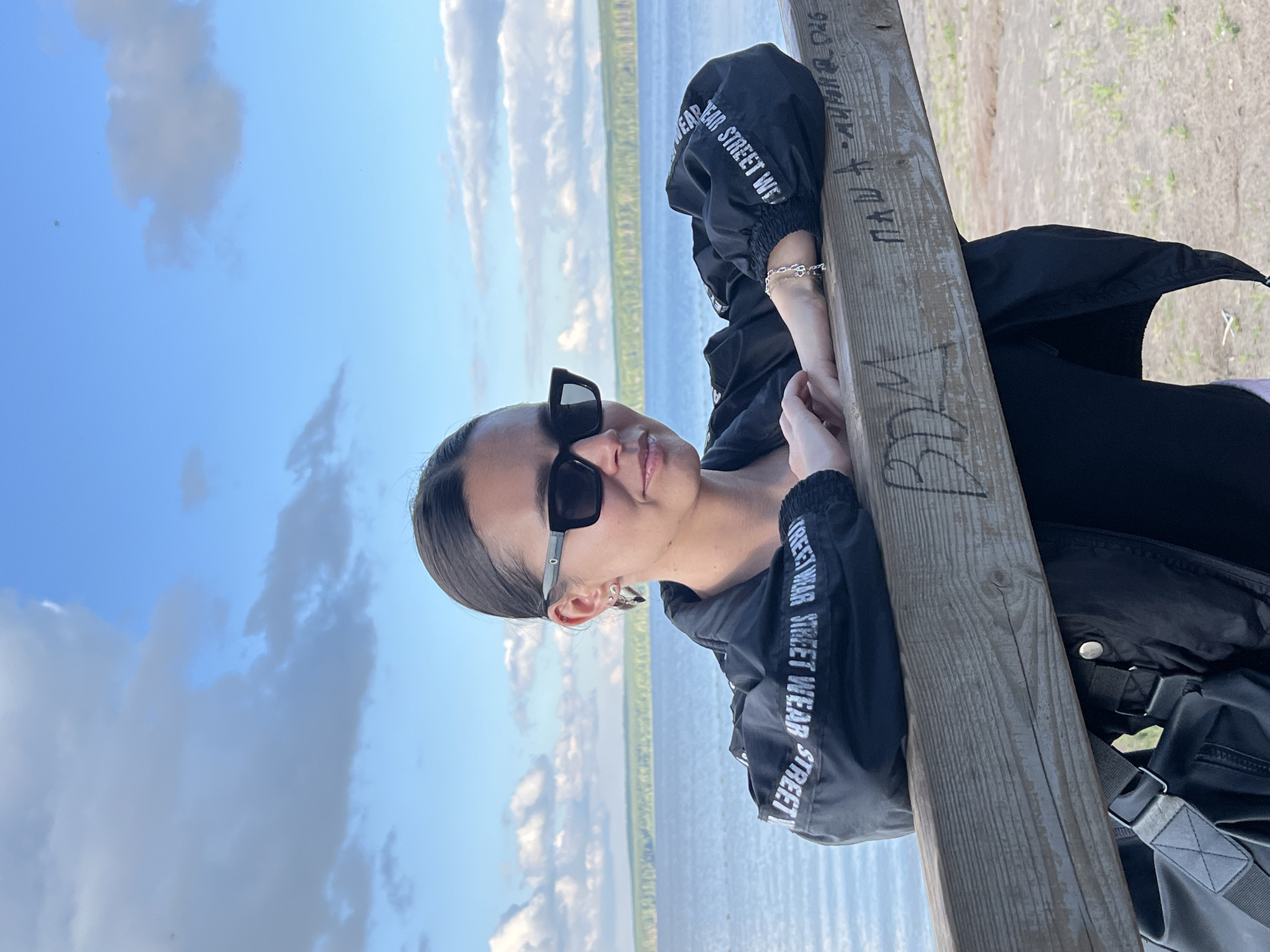 A young woman wearing sunglasses and a black jacket with white text on the sleeves is leaning against a wooden post. The text on her jacket reads "STREET WEAR STREET WEAR STREET WEAR". She has dark hair pulled back and is looking towards the camera with a slight smile. Her right arm is resting on the post, and she is wearing a silver bracelet.
Behind her, a body of water, likely a lake or river, stretches towards a distant green shoreline under a bright blue sky with scattered white clouds. The ground around the post appears to be sandy and covered with sparse dry grass.
There are some markings carved into the wooden post. One marking appears to be a set of initials or a name, possibly "PDK". Another set of markings, in a script-like font, is also visible. The lighting suggests it is daytime, likely a sunny afternoon, given the clear sky and shadows. The overall mood appears relaxed and casual. The location is identified as Chaikovsky, Russia.