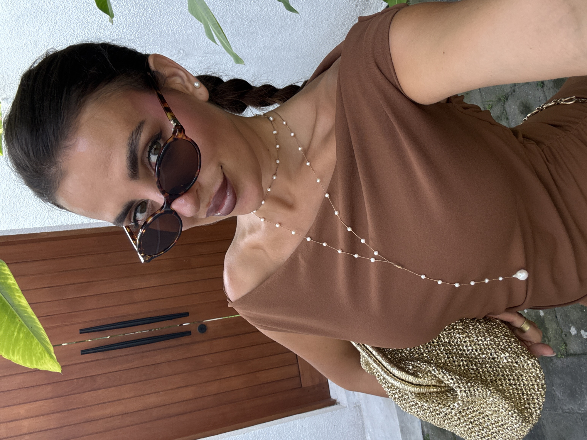 A woman with brown hair in braids and wearing sunglasses and a brown off-the-shoulder dress is taking a selfie outdoors in North Kuta, Indonesia. She has a pearl necklace and is holding a gold woven bag. In the background, there is a wooden door and green foliage. The ground appears to be concrete with some vegetation growing nearby. The image is taken at an angle, tilting slightly to the left.