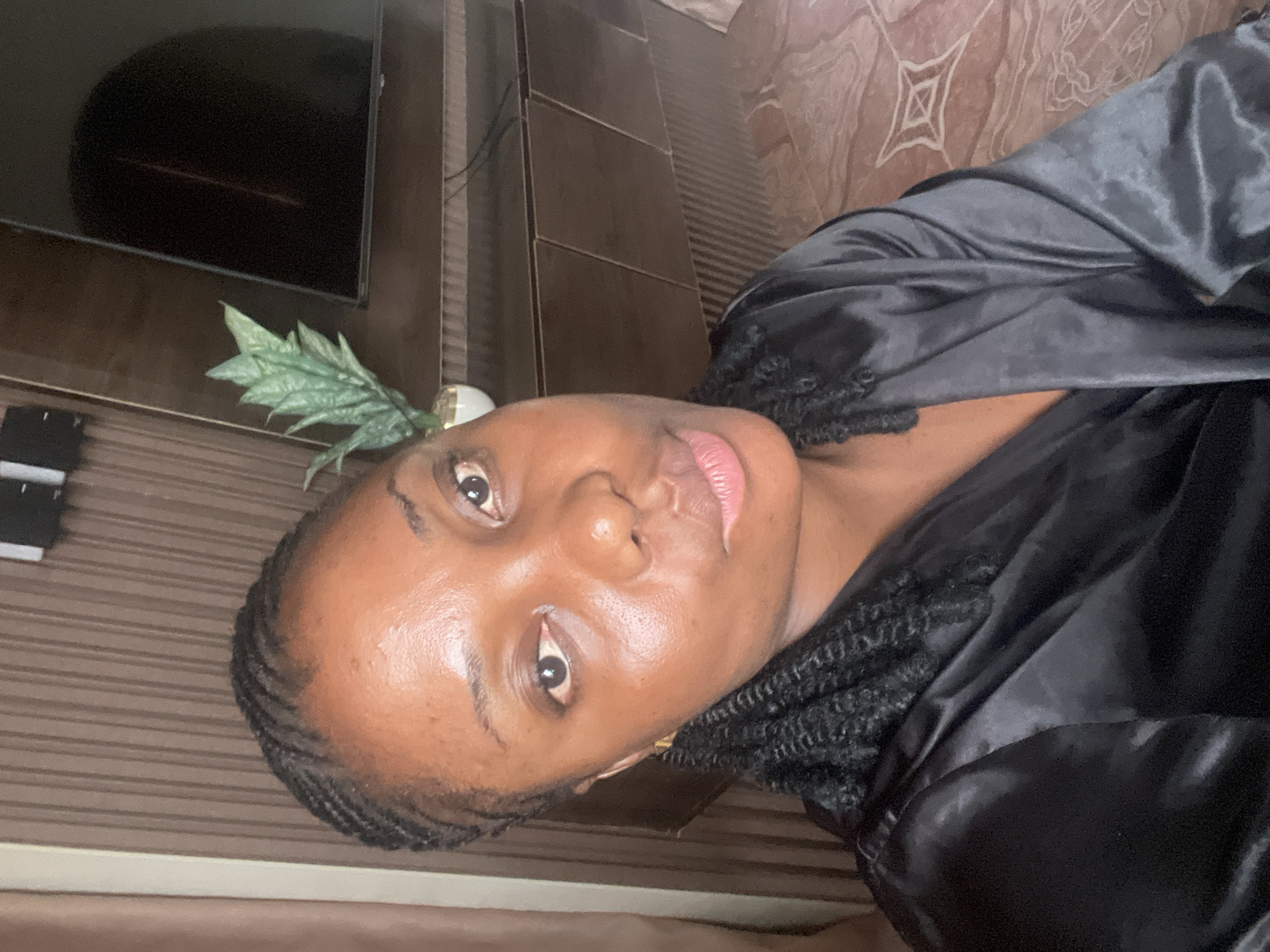 A dark-skinned person with braided hair, wearing a black satin robe, is pictured indoors against a background of brown-toned walls, possibly in Jos, Nigeria. A flat-screen television and a decorative plant are visible in the background. The person's face is in focus, and they are looking directly at the camera.