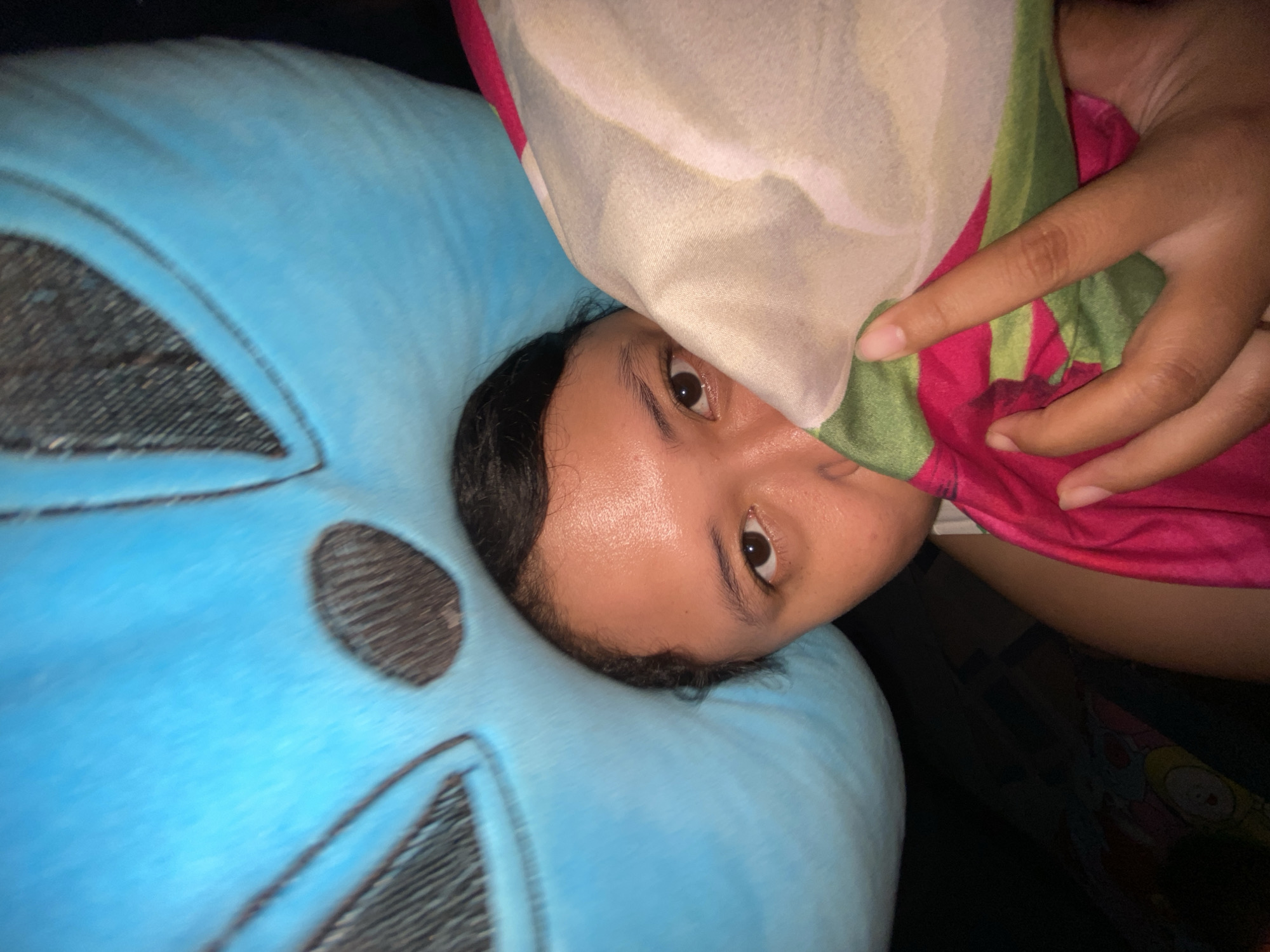 This close-up, angled image, taken in Gelam, Indonesia, captures a person resting indoors, likely in dim lighting. The subject, appearing to be young, is partially covered by a colorful blanket or sheet, peeking out with one eye directly at the camera. Their face, including the visible right eye, forehead, and cheek, has a warm skin tone, and there is a noticeable sheen on the forehead, possibly from sweat. Dark hair frames the face.
The person's head rests on a large, soft-looking blue object, which appears to be a pillow or cushion. This blue item features faint black outlines, suggesting a stylized design, possibly abstract facial features or a cartoon character, with textured dark areas within the blue fabric.
A vibrant piece of fabric, predominantly white or cream with bold green and magenta patterns, covers much of the person's upper body and obscures part of their face. A hand, with fingers gently curved, is visible near the top right of the frame, seemingly holding or adjusting this patterned textile. The background beyond the person and pillow is dark and indistinct, hinting at other soft furnishings or a dark environment. The scene conveys a quiet, personal moment, possibly either waking up or settling down for rest. There is no visible text in the image.