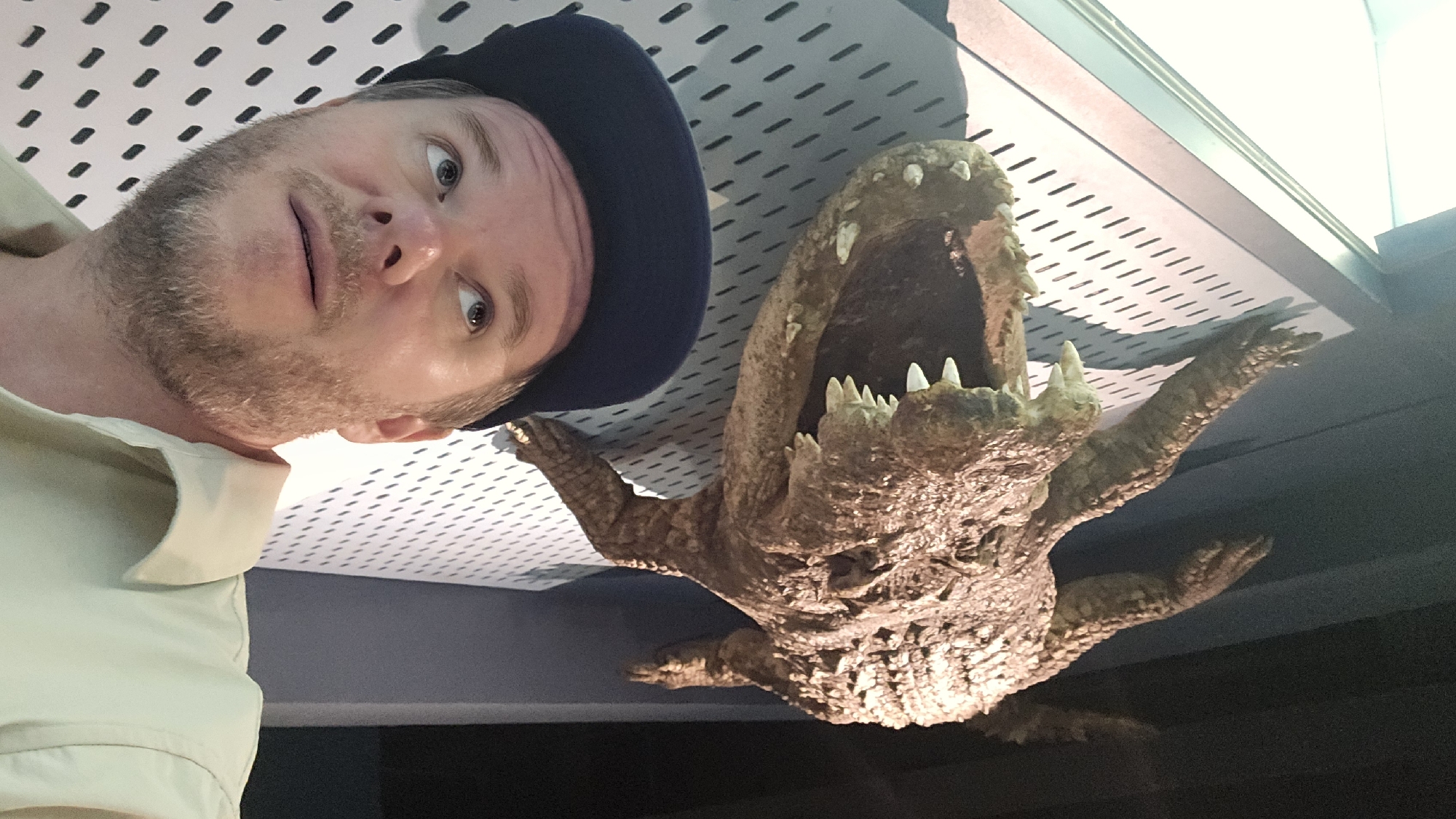 In Gongshu District, China, a man wearing a dark blue baseball cap and a light-colored collared shirt is looking upwards, his face close to the camera. Behind him, suspended on a perforated white wall, is a large, reptilian head with its mouth open, revealing sharp teeth. The creature appears to be a prop or an exhibit, with textured brown skin. The lighting casts shadows, highlighting the details of both the man and the prop.