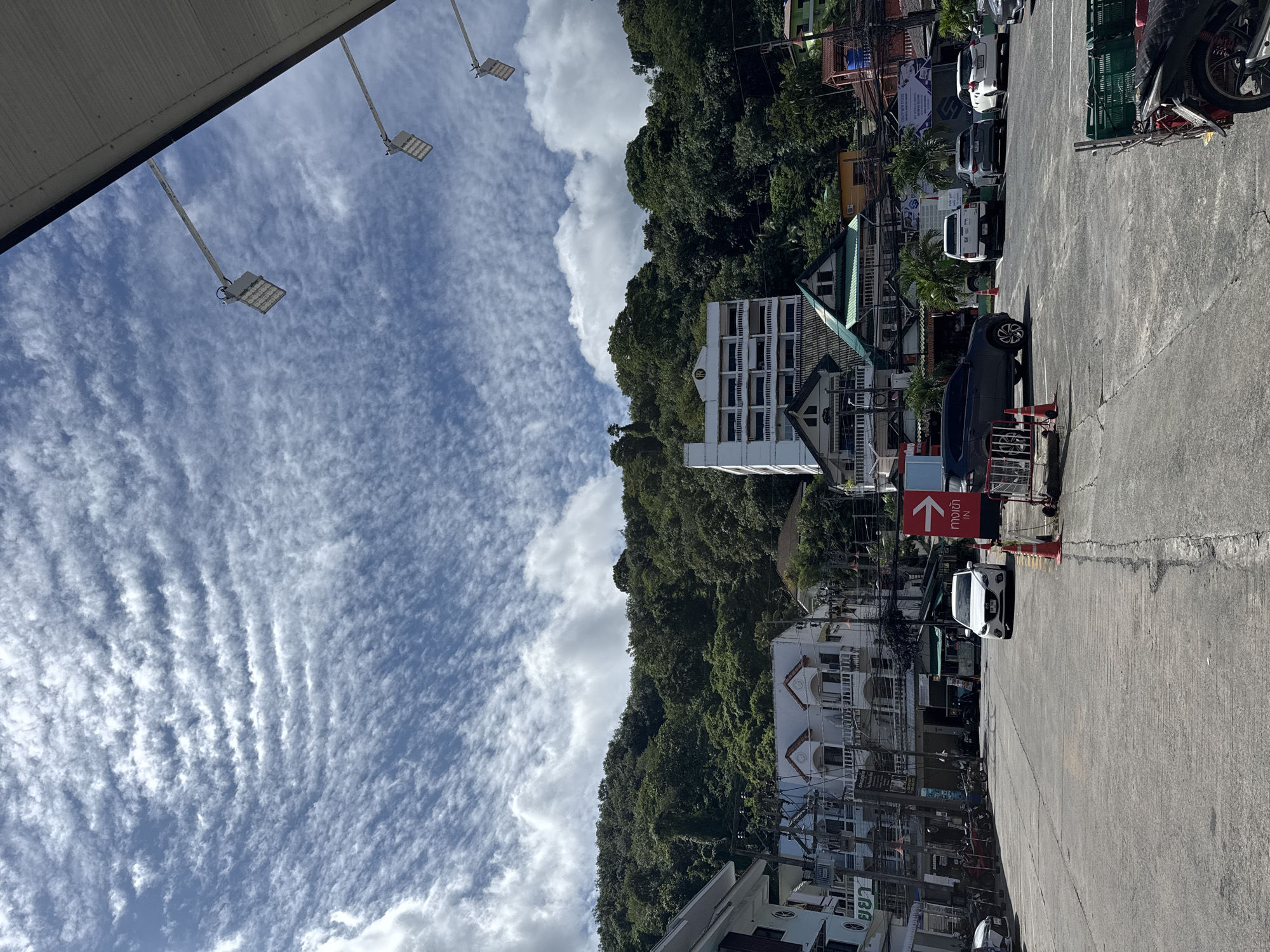 The image depicts a street scene in Patong, Thailand, with a focus on a paved road leading towards a dense, tree-covered hillside under a sky filled with wispy clouds. Several buildings of varying architectural styles are visible along the street, ranging from multi-story structures with balconies to smaller, traditional-looking houses with pitched roofs. Some of these buildings appear to be commercial establishments, with visible signage. A white car is parked in the middle of the road, and several other vehicles, including SUVs and motorcycles, are parked on the sides or in designated parking areas. The street is lined with traffic cones and bollards, suggesting a controlled environment, possibly a parking lot or a street with traffic management. Overhead, industrial-looking lights are mounted on poles, and electrical wires are strung between buildings, indicating an urban setting. The overall atmosphere is that of a bright, sunny day in a bustling Southeast Asian town.