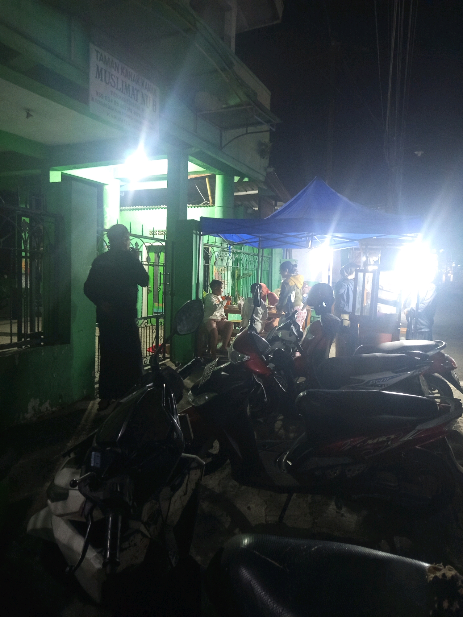 The image depicts an outdoor scene at night in Singosari, Indonesia, in front of a building with green walls and a sign that reads "TAMAN KANAK-KANAK MUSLIMAT NU 8". Several motorcycles are parked in the foreground, some of which are partially obscured by darkness and glare. In the background, a blue tent is set up, illuminated by bright lights, and several people are gathered underneath it. One person in the foreground is standing near a green gate and appears to be smoking. Another person is sitting on a chair under the tent, and others are visible in the background, some appearing to be engaged in conversation or activity. The overall impression is that of a lively street scene with temporary food stalls or gathering points operating at night.