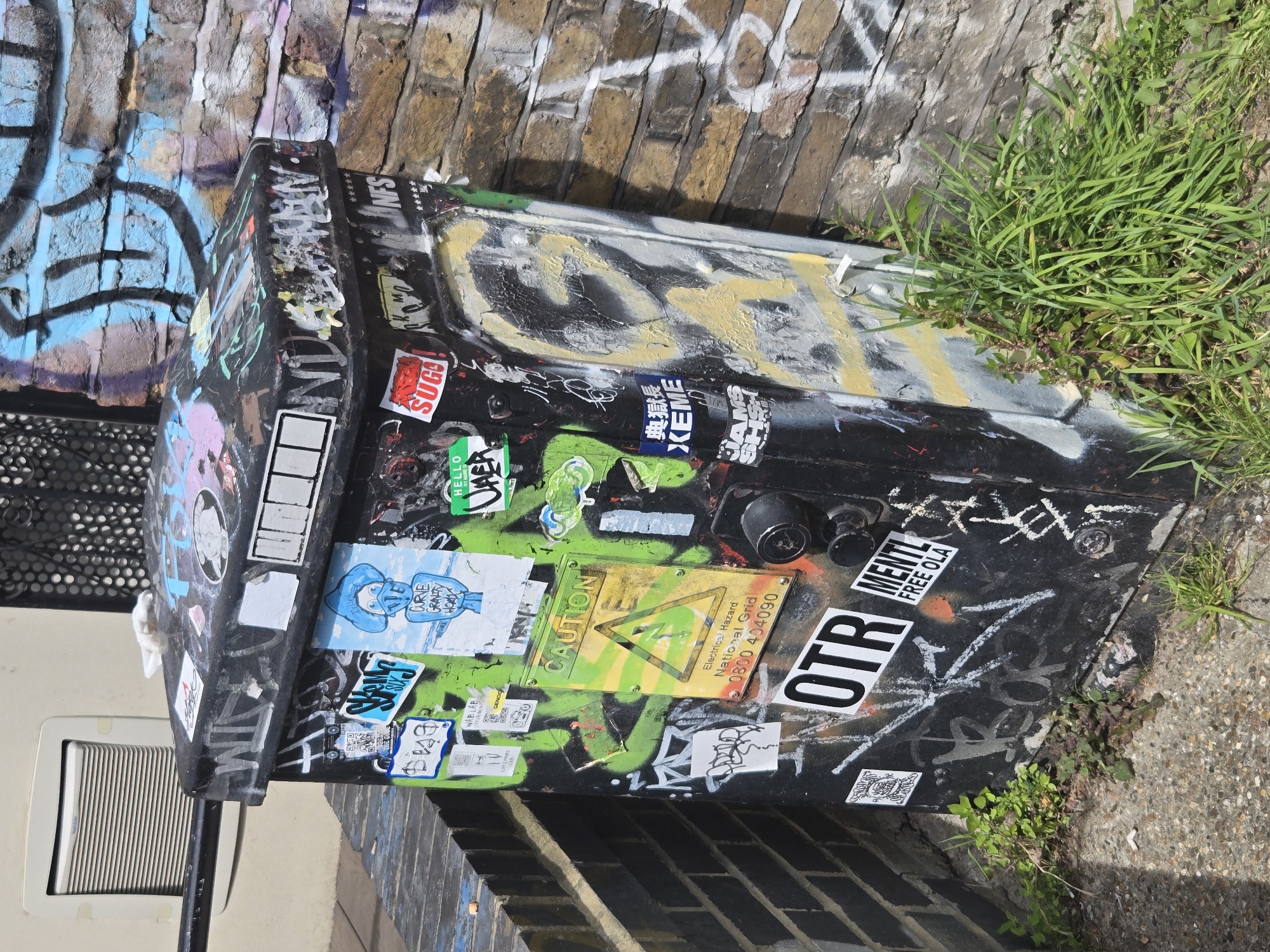 The image shows a black electrical box, covered in graffiti and stickers, located in London, United Kingdom. The box sits adjacent to a brick wall with additional graffiti. Patches of grass and weeds are growing around the base of the box. A wall vent is visible to the left of the box. The stickers include labels such as "CAUTION", "SUGO!", "XEME", "OTR" and "MENTAL FREE OLA". The box has a latch, and a sign that reads "Electrical Hazard National Grid 0800 404090"
