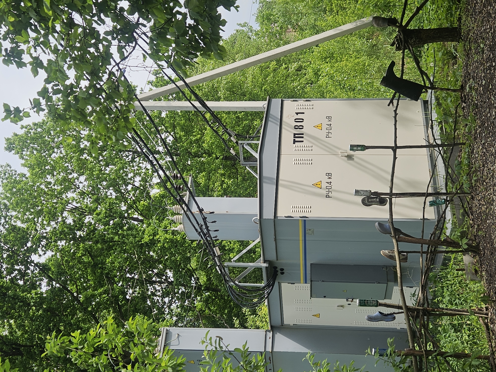 The image depicts an electrical substation in a wooded area. The substation is composed of metal boxes, some painted light gray and blue, with warning signs in Russian. Power lines connect to the substation from above. In the foreground, a makeshift fence constructed from sticks is adorned with discarded shoes and cans. The location is identified as Посёлок комбината стройматериалов-1, Russia. The environment consists of trees and undergrowth, suggesting a rural or suburban setting.