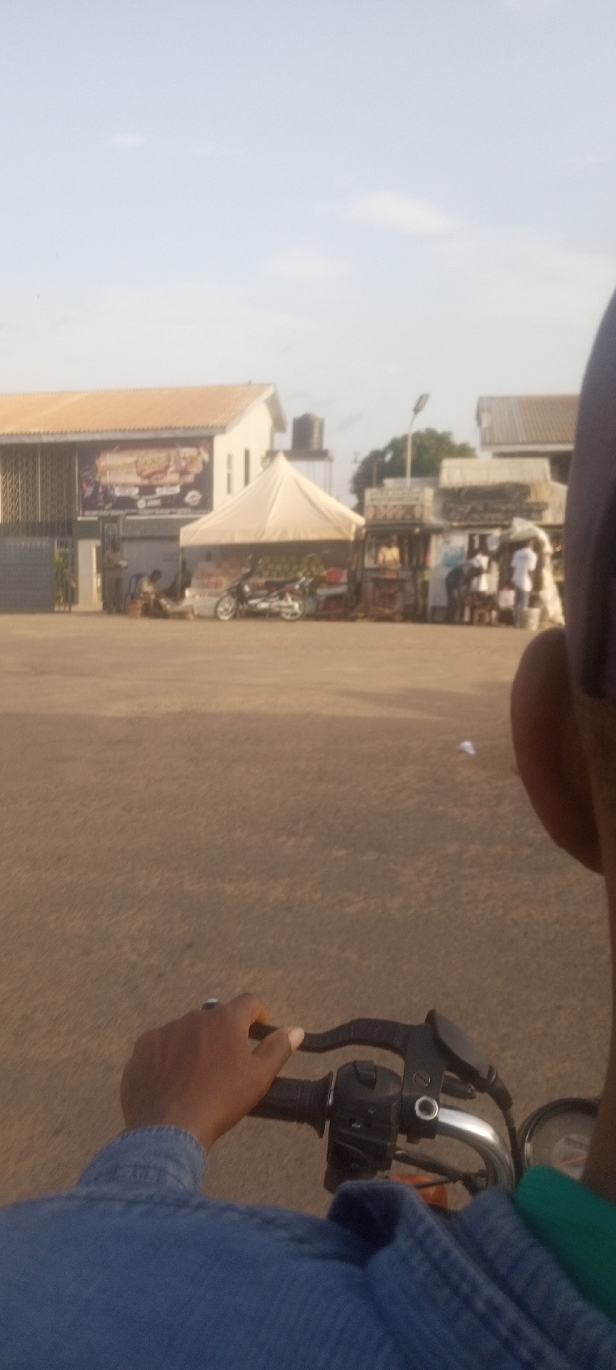 A ground-level point-of-view shot from what appears to be a moving motorcycle, showing a road surface ahead and what looks to be an outdoor market scene. In the foreground, the motorcycle handlebars and controls are in focus, along with a portion of the rider's arm and clothing. The road surface appears to be compacted dirt. In the background, a cluster of commercial buildings and temporary market stalls are visible. There are people visible near the stalls, engaging in activity that could include buying or selling goods. A white tent-like structure stands between the buildings and the market activity, with another motorcycle parked alongside it. The buildings in the background include a two-story structure with a billboard or large advertisement, as well as other single-story structures. A water tower is visible on the roof of the two story structure. The sky is light and cloudless. The location is Narayi, Nigeria.