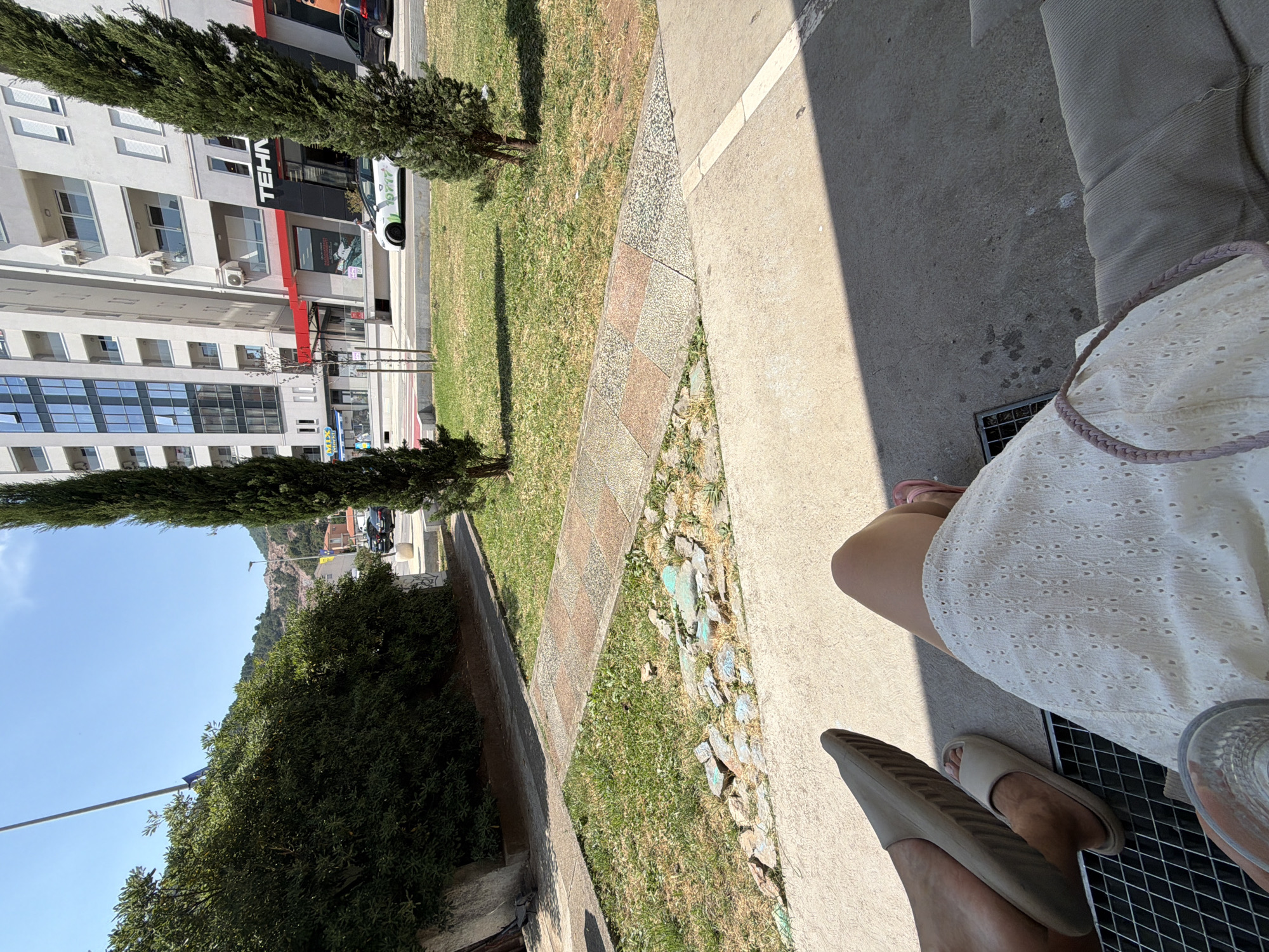 A low-angle shot captures a sunny outdoor scene, likely in Budva, Montenegro, based on the surrounding architecture and vegetation. In the foreground, a person's legs are visible, wearing a white eyelet dress and light-colored slide sandals. Another person is wearing darker slide sandals. The ground is concrete, with a metal grate visible near the feet. A shadow from an unseen structure falls across the ground.
To the left, a tall, slender cypress tree stands in front of a modern apartment building with multiple floors and balconies. Some windows are visible, and air conditioning units are mounted on the building's facade. Text on the building includes "TEHN" in a black sign with a red stripe below it. Further down the street, other buildings and cars are visible.
The middle ground features a patch of grass with a decorative stone pathway. Beyond that, more lush green vegetation and a tree can be seen. The background shows a hillside covered in greenery and some buildings, indicating a hilly coastal environment. The sky is a clear, bright blue, suggesting a pleasant, sunny day. The overall mood is relaxed and casual, hinting at a moment of rest or enjoyment outdoors.