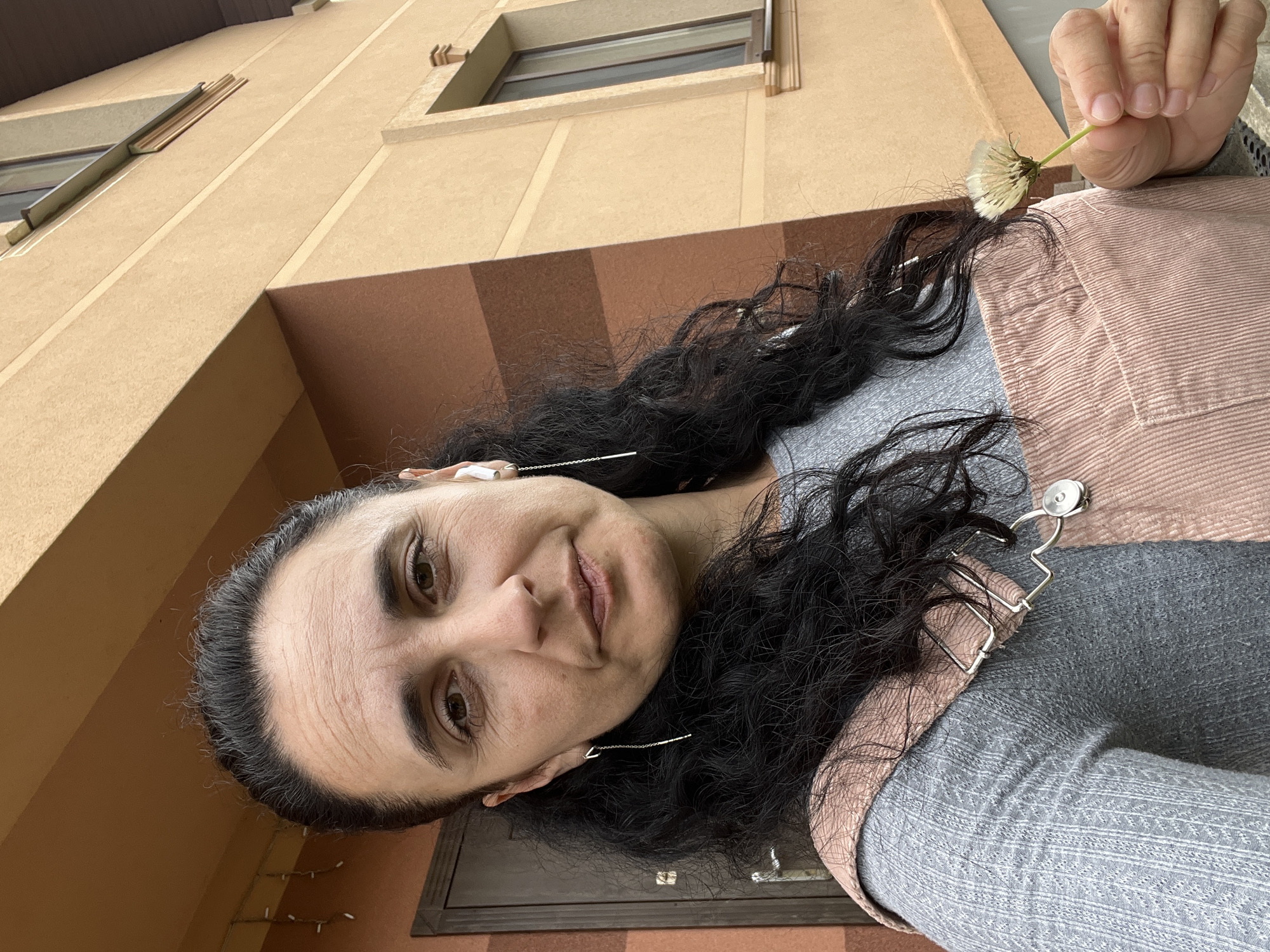 A woman with long, dark, curly hair is prominently featured in a close-up outdoor portrait, holding a fluffy white dandelion puff in her right hand. She looks directly at the viewer with a subtle, pleasant expression, her eyes wide and a hint of a smile on her lips. She wears a grey long-sleeved top layered under a light pink or peach-colored corduroy-textured jacket or vest, which features a distinctive silver, abstract-shaped brooch or clasp near her shoulder. Long, delicate silver chain earrings hang from her ears.
The background consists of a multi-story building with a light beige or tan facade, featuring horizontal brown striped sections and rectangular windows. The warm, even lighting suggests it is daytime. Below what would be ground level or a lower section of the building, a string of small, white lights is visible. The image appears to be taken from a slightly low angle, looking up towards the woman and the building.
The setting is Onokivtsi, Ukraine. No visible text is present in the image.