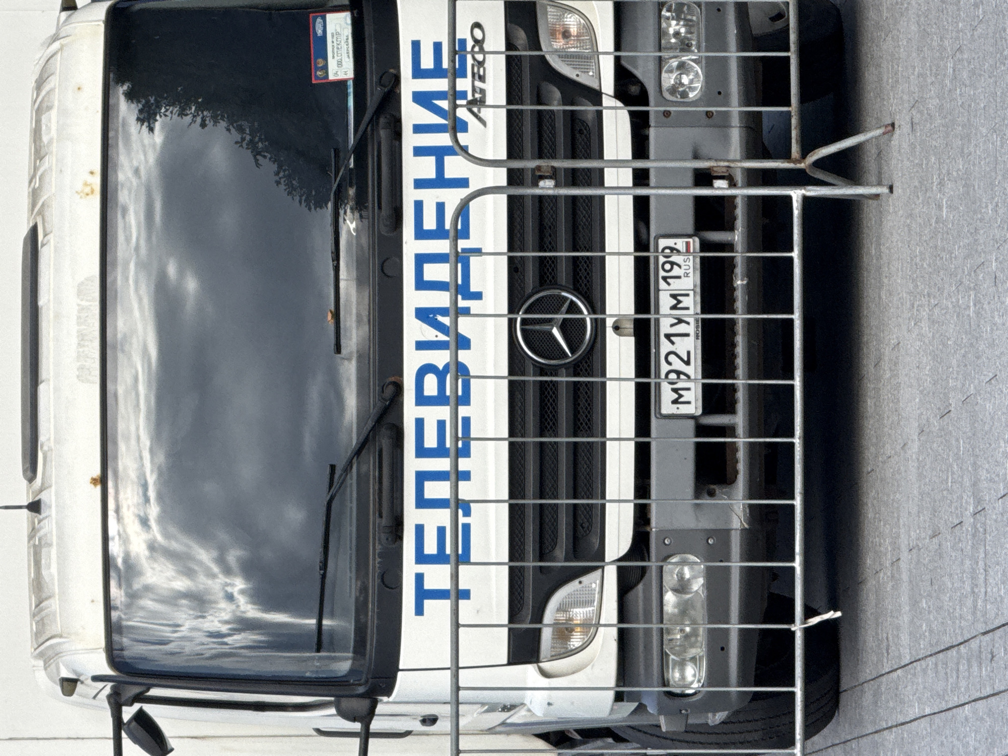 A white Mercedes-Benz Atego truck is parked behind a metal barrier, in a street in Moscow, Russia. The truck has the word "ТЕЛЕВИДЕНИЕ" (Television) written in blue letters on its front. The license plate reads "M921YM 199". The truck's windshield reflects clouds and trees. The street pavement is visible in the foreground.