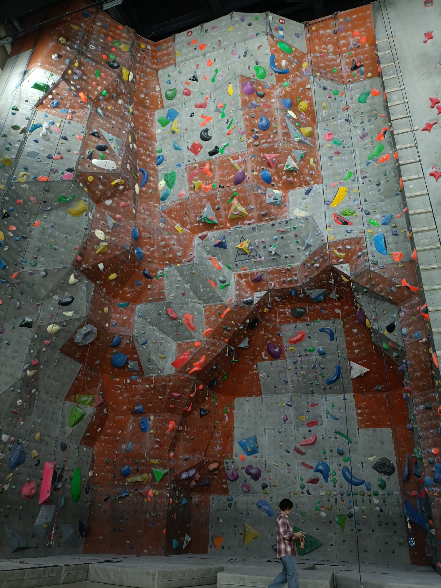 The image depicts a climbing wall at the "Rock-Zone" climbing center, located in Krasnoyarsk, Russia. The wall is tall, complex, and densely populated with colorful climbing holds. The surface is composed of both gray and reddish-brown panels. Ropes hang down from the top, indicating climbing routes. A metal ladder runs along the right side of the wall. In the foreground, a person dressed in a red and white checkered shirt and jeans is walking. The scene appears to be indoors, with artificial lighting and a padded floor beneath the climbing wall.
