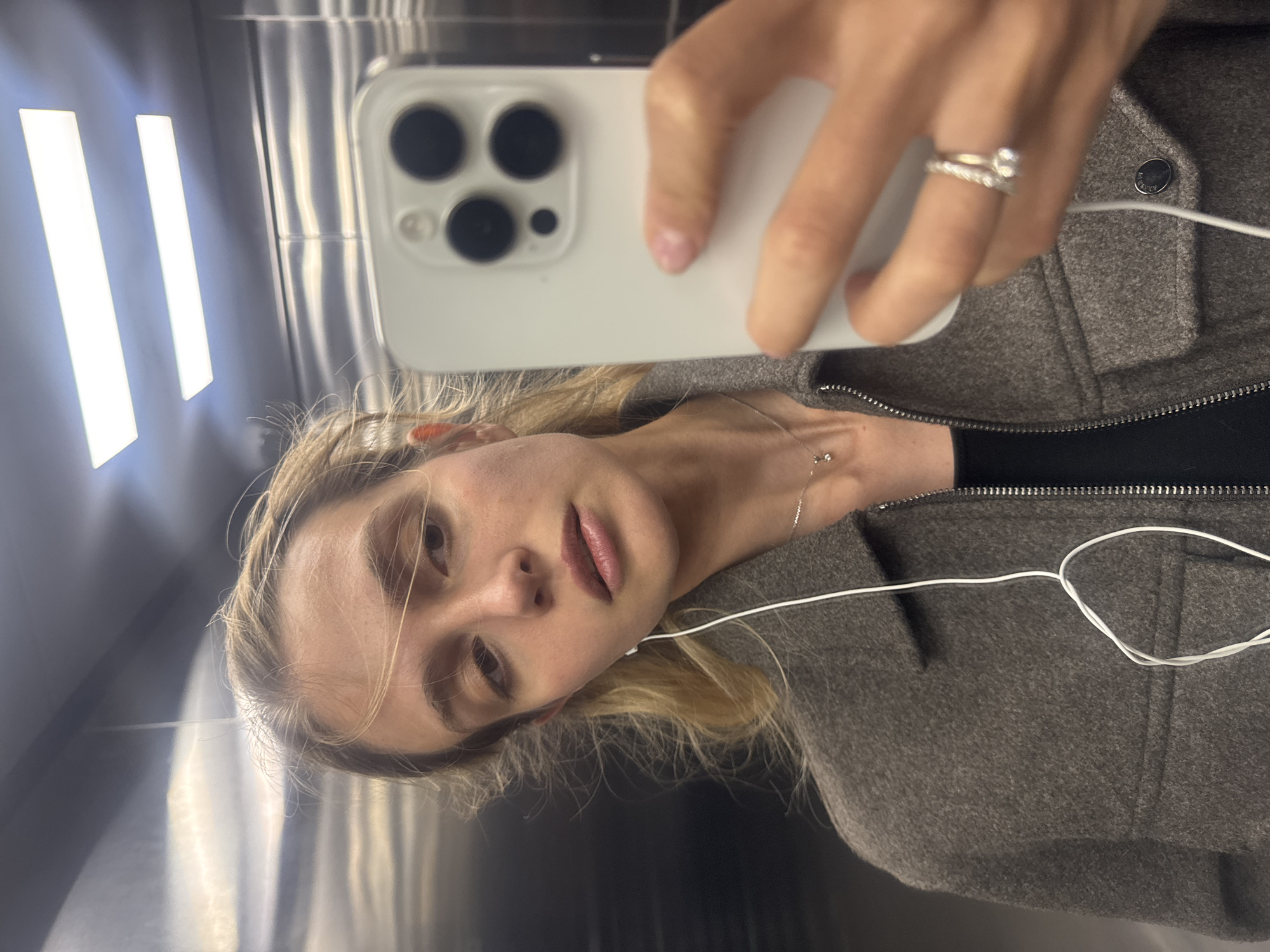 A young woman with blonde hair takes a selfie in a reflective surface, likely a mirror or polished wall, capturing her reflection and the white smartphone she holds. She is looking directly towards the camera with a neutral to slightly pensive expression, her lips slightly parted. Her right hand, adorned with a ring featuring a clear stone, holds the white smartphone which clearly shows a triple-lens camera system on its back.
She is wearing a dark gray or brown textured jacket or coat with a visible zipper and a single button detail. White wired earphones are in her left ear, with the wire extending across her chest and disappearing behind her jacket. A delicate silver chain necklace with a small pendant is visible on her neck. The background features bright, vertical light strips reflecting in the shiny surface, suggesting an indoor, possibly metallic, environment such as an elevator or a modern interior. There is no visible text in the image.