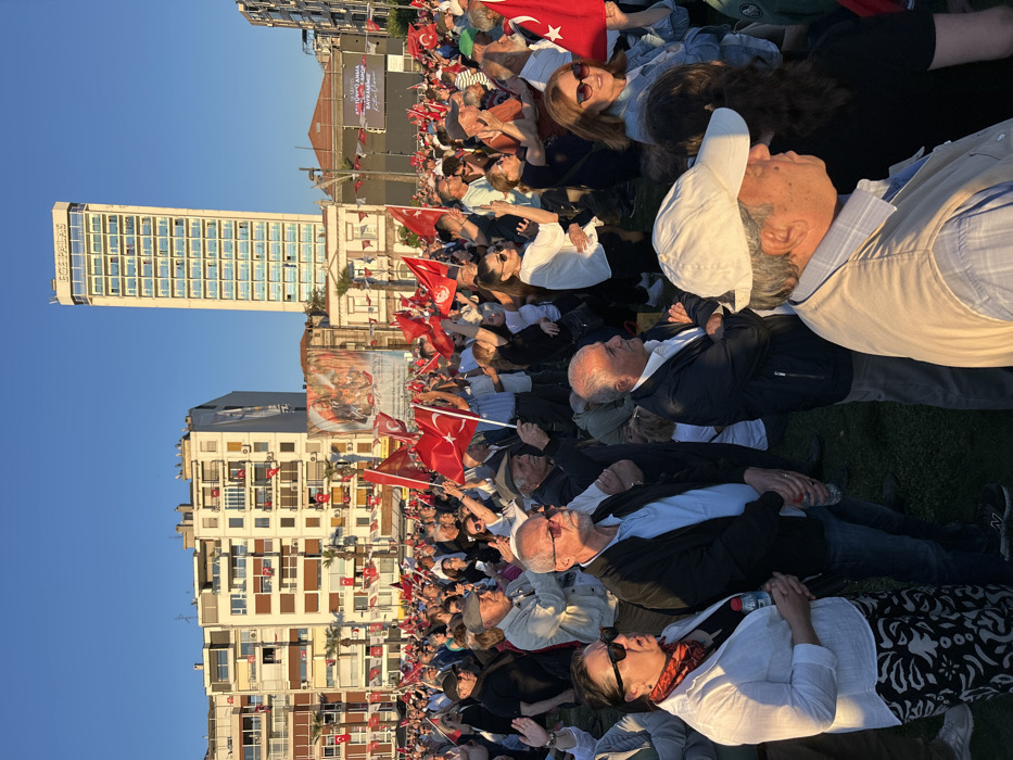 A large crowd of people, mostly adults, are gathered in an open area in İzmir, Türkiye, at the coordinates 38.436 latitude, 27.14 longitude. The crowd is standing on a grassy patch of land surrounded by buildings, including a tall building with "BOS PHALAS" written on it. The people are holding Turkish flags, with some people waving them. The crowd is facing a stage, and people are standing on the stage holding up banners. The people in the crowd are looking towards the stage, some are talking to each other, and some are waving their flags. The sky is blue and the sun is shining. The scene appears to be an outdoor political rally or demonstration.