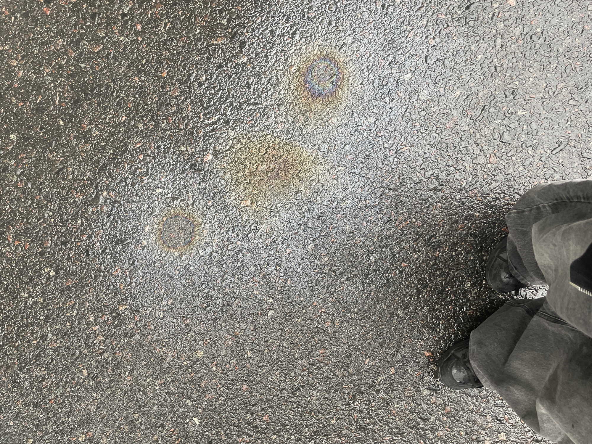 The image shows a section of asphalt pavement, likely a street or parking area. The pavement is wet, and there are three circular stains with iridescent sheens, suggesting the presence of spilled oil or similar petroleum-based liquid. The colors of the iridescent sheens range from blues and purples to yellows and oranges. To the right of the frame, a person's legs are visible, clad in grey pants and dark-colored shoes. The environment is urban, with a focus on the pavement surface. There is no visual information to determine if this photo was taken in the city of Buenos Aires, Argentina.