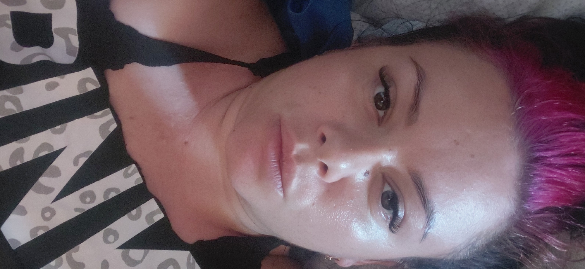 This is a close-up, head-and-shoulders selfie of a woman in what appears to be a bedroom setting. The woman has dark hair with bright pink highlights. She is wearing a black top with a leopard print pattern on the front that says "PINK". The woman has dramatic false eyelashes and appears to have some makeup on, including lip gloss. Her skin has a dewy or oily sheen to it. The background is soft and out of focus, with a hint of a grey textured surface behind her head and a blue object to her left. There is a small gold hoop earring visible in her right ear. The image was taken in Marynivka, Ukraine.