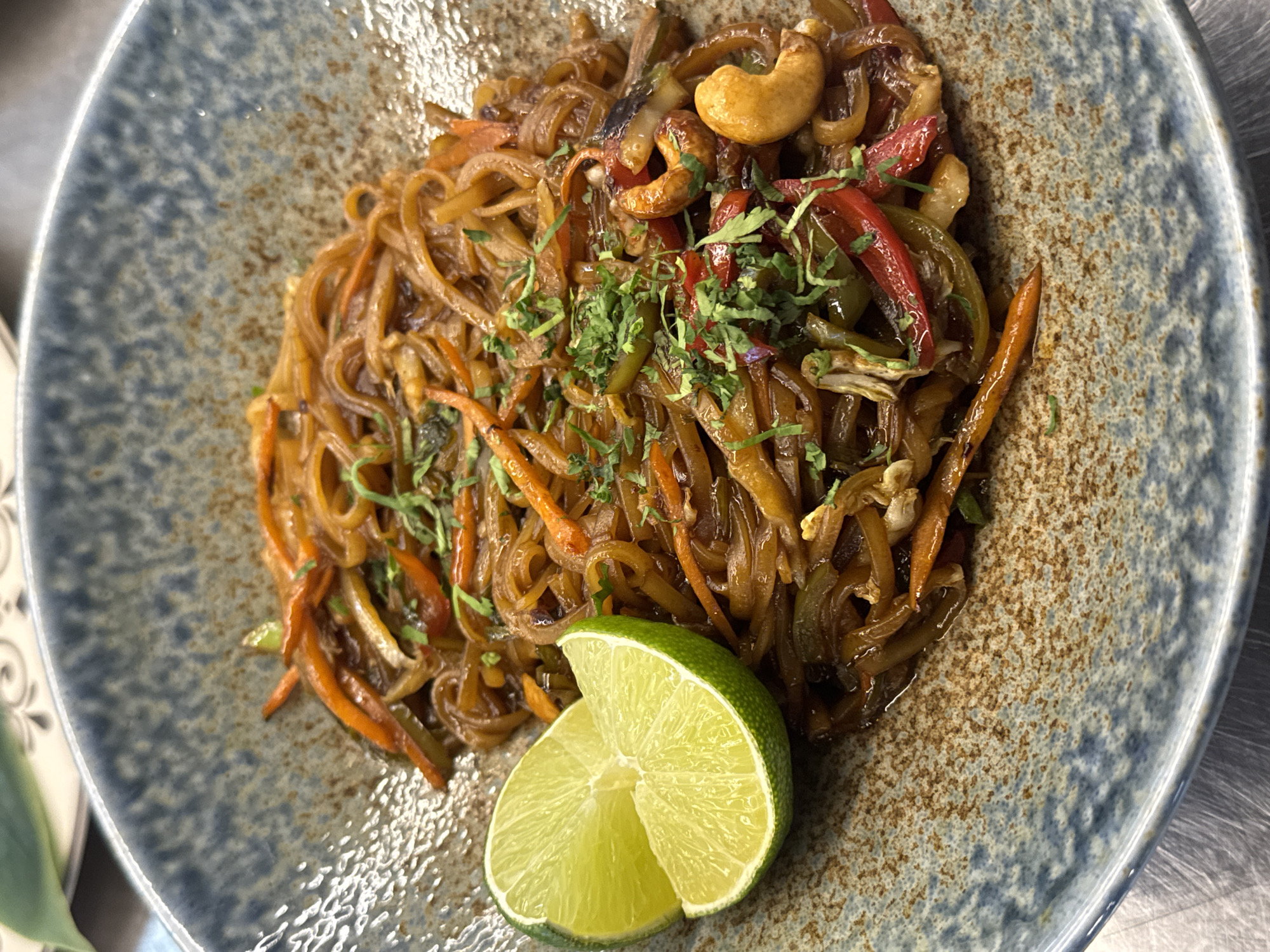 The image shows a close-up of a bowl of stir-fried noodles with vegetables and cashews. The dish is served in a rustic, speckled blue and brown ceramic bowl. A wedge of lime is placed on the left side of the bowl. The noodles appear to be thin and dark brown, mixed with julienned carrots, red and green bell peppers, and whole cashews. Chopped fresh green herbs are sprinkled on top. The background is blurred, but it suggests an indoor setting, possibly a restaurant. The lighting is natural and highlights the textures and colors of the food. The image does not contain any information that would allow it to be placed in Sofia, Bulgaria.
