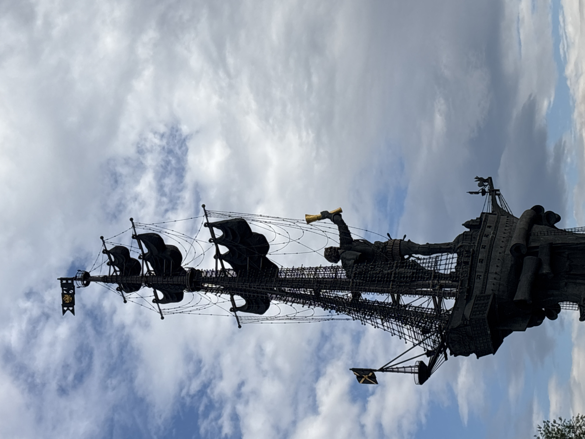 The image shows a large, dark-colored monument depicting a ship with figures, viewed from a low angle against a cloudy sky. The ship features detailed rigging, sails, and several human figures in various poses, including one holding a golden-colored trumpet. The location is not possible to specify because the city is unknown.