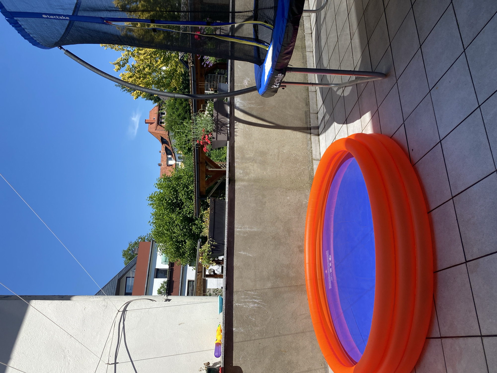 This media file captures an outdoor scene on a sunny day, likely in a residential backyard or patio. The primary objects in focus are an orange inflatable pool with a blue interior, positioned on a tiled surface, and a large blue trampoline, part of which is visible in the upper left quadrant of the image. The trampoline has a black safety net and is supported by metal poles.
In the background, a residential neighborhood can be seen with houses featuring red-tiled roofs and green trees. The sky is a clear, vibrant blue, suggesting fair weather and a time of day that is likely mid-morning to afternoon due to the bright sunlight and the shadows cast by the trampoline. The scene evokes a sense of summer leisure and outdoor fun, typical for warm weather activities.
There is some visible text on the trampoline net that reads "tectake". The overall impression is one of a bright, cheerful day, prepared for outdoor recreation.