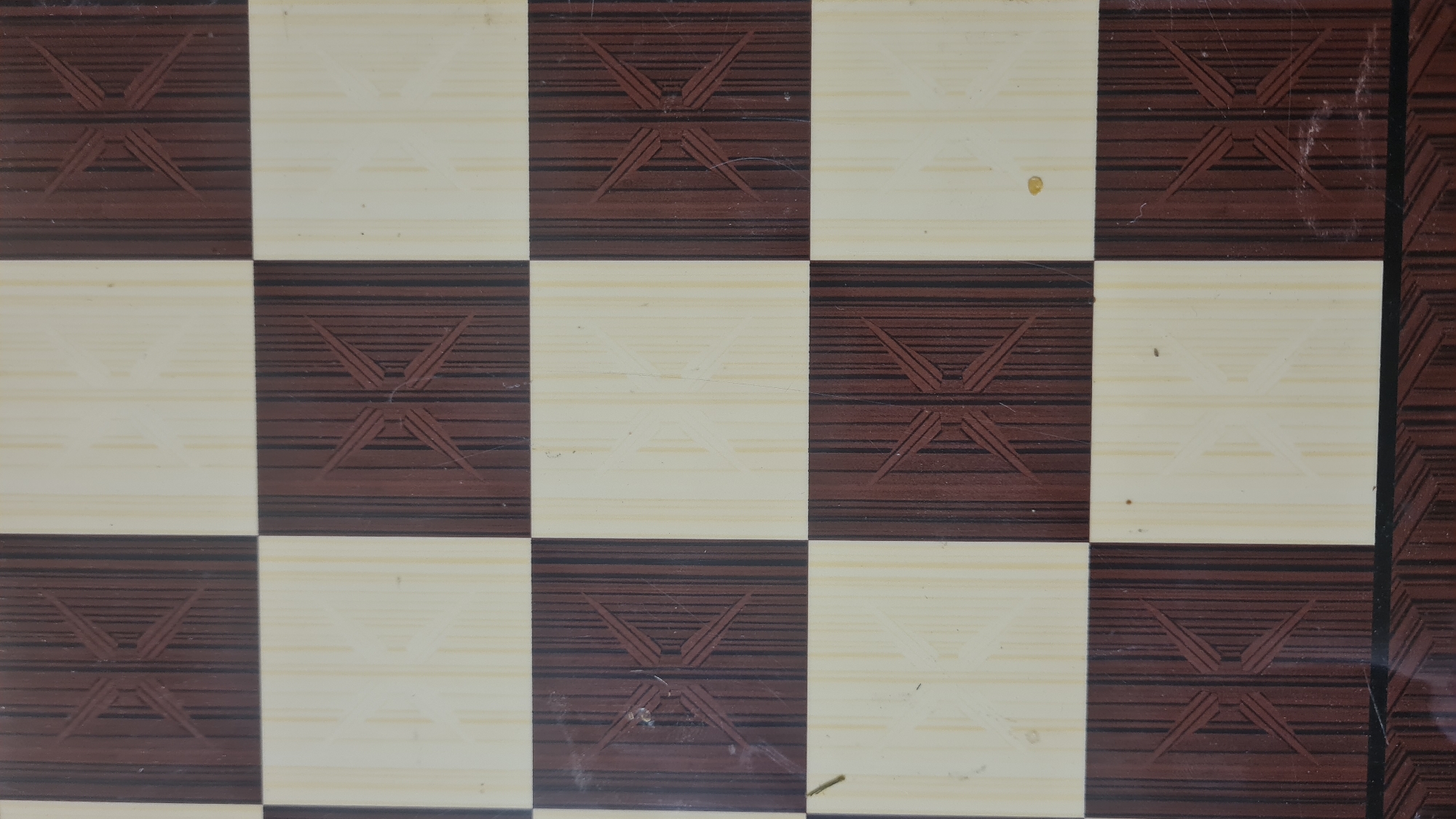 The image presents a close-up, overhead view of a checkerboard pattern, consistent with a chessboard or a similar game board, located in the Lachish Regional Council, Israel. The board is composed of alternating dark brown and light cream/beige squares. Each individual square features a subtle, embossed or printed "X" or star-like motif. On the dark brown squares, this pattern is rendered in a slightly lighter shade of brown, while on the light cream squares, it appears in a darker cream tone, creating a distinct yet understated contrast.
The surface of the board exhibits a slight sheen, suggesting a laminate or plastic coating. Visible notable details include minor scratches, scuff marks, and dust particles scattered across the squares. For instance, a small yellowish spot is noticeable on one of the light squares in the upper right quadrant, and several faint linear scratches crisscross various squares. Both the dark and light squares feature a printed horizontal wood grain texture. No game pieces are present, and therefore no activity is depicted. There is no visible text on the board. The lighting is even, indicating an indoor setting.