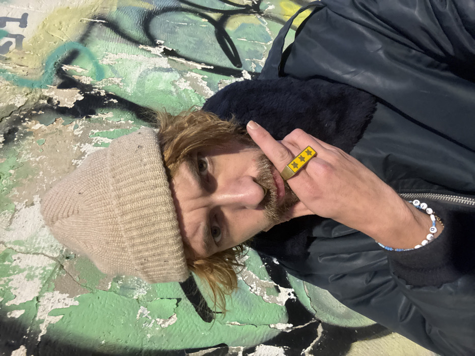 A light-skinned person with shoulder-length blond hair and a beard wears a beige beanie and a dark jacket with a fur collar. They are positioned against a graffiti-covered wall. The person is flipping off the camera, and a wide gold ring with yellow stars adorns their middle finger. A beaded bracelet is visible on their wrist.