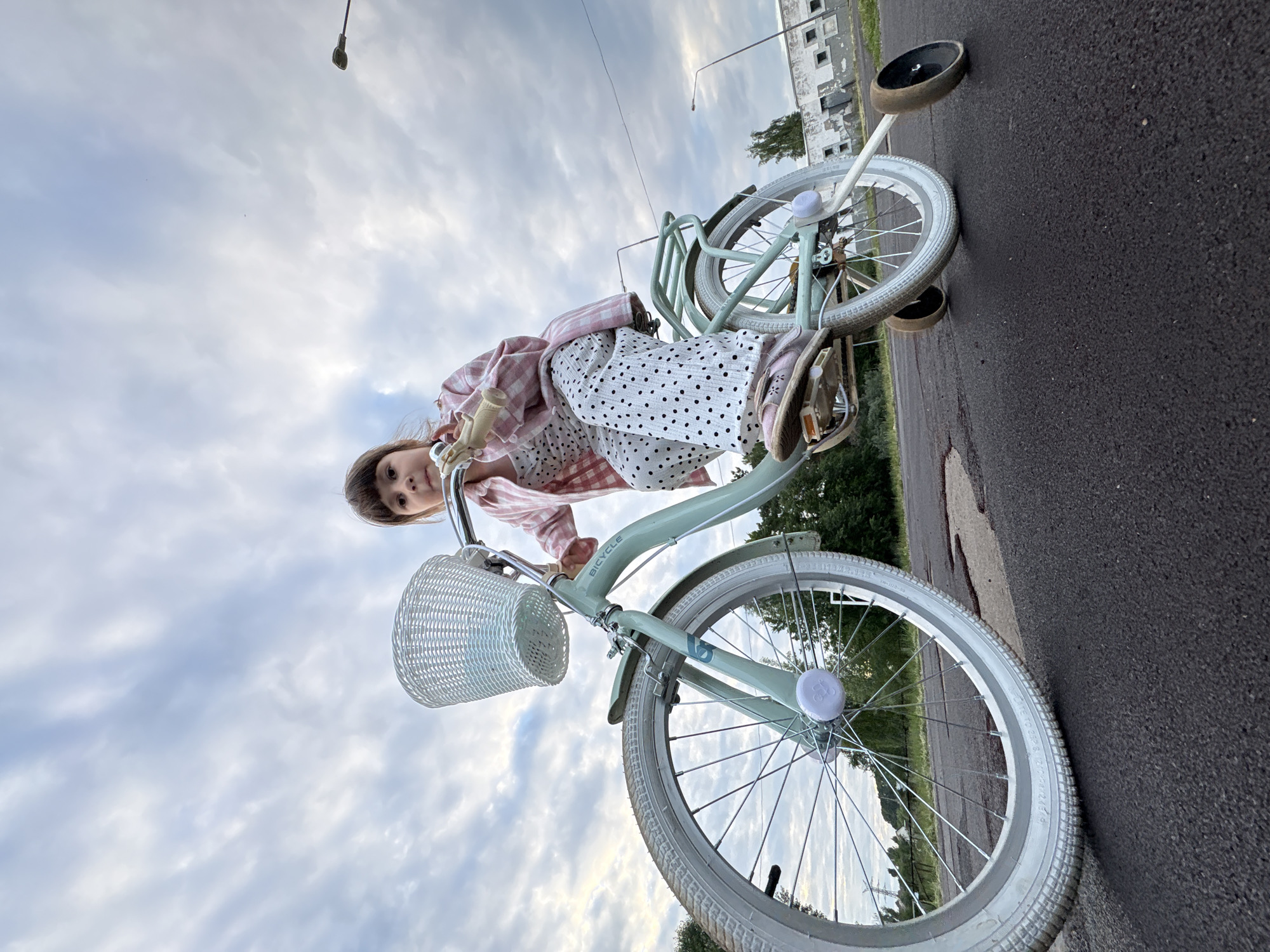 An outdoor, low-angle, wide shot captures a young girl, seen from the chest down, sitting on a light teal bicycle with training wheels. The girl wears a pink and white checkered jacket over a white polka-dot jumpsuit and pink shoes. The bicycle has a white wicker basket attached to the handlebars and a white reflector on the front wheel. The scene takes place on an asphalt surface, possibly a road or pathway, with some dark stains and cracks visible. In the background, there are trees, a power line tower, and a weathered, long building with multiple windows. The sky is overcast with a mix of gray and white clouds, and a streetlamp is visible in the upper right corner. The image appears to be taken in the city of Chaikovsky, Russia, based on its visual characteristics typical of such a location.