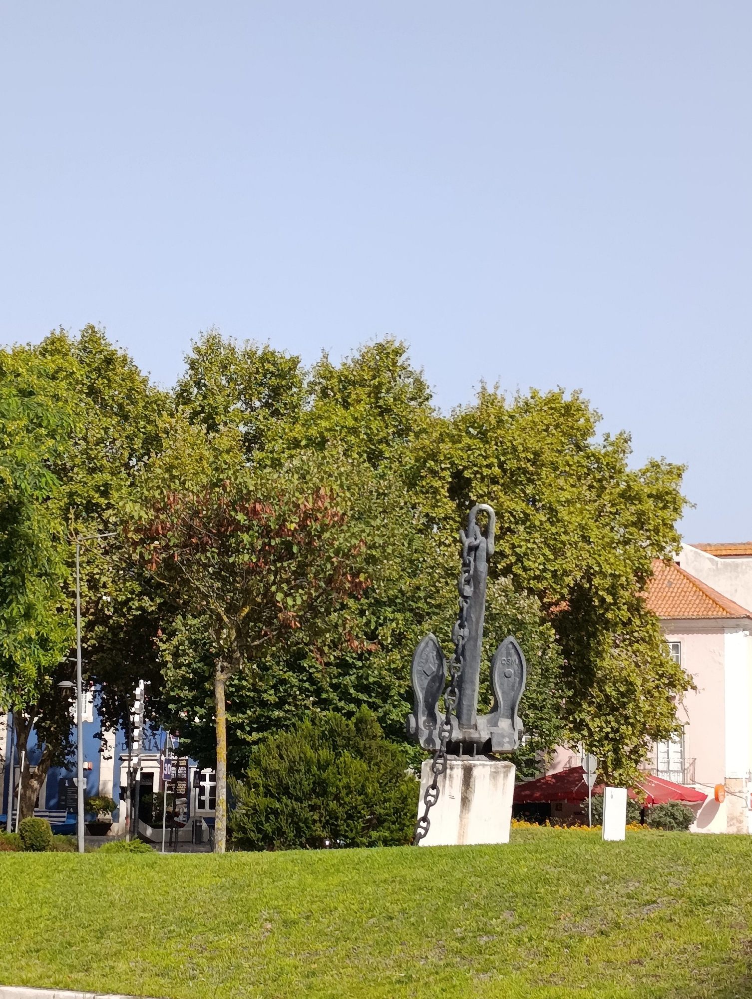 The image depicts a large metal anchor monument situated on a grassy mound in the city of Setúbal, Portugal. The anchor is dark grey and appears to be made of metal, with a prominent chain attached to its upper part. It stands on a light-colored, possibly concrete, pedestal. Behind the monument, a line of lush green trees forms a backdrop. To the right, a light pink building with a terracotta tiled roof is visible, and to the left, a blue building is partially obscured by the trees. In the distance, red awnings can be seen, suggesting a commercial area. The sky is clear and pale blue. The overall scene suggests an outdoor public space, possibly a park or a plaza, on a clear day.