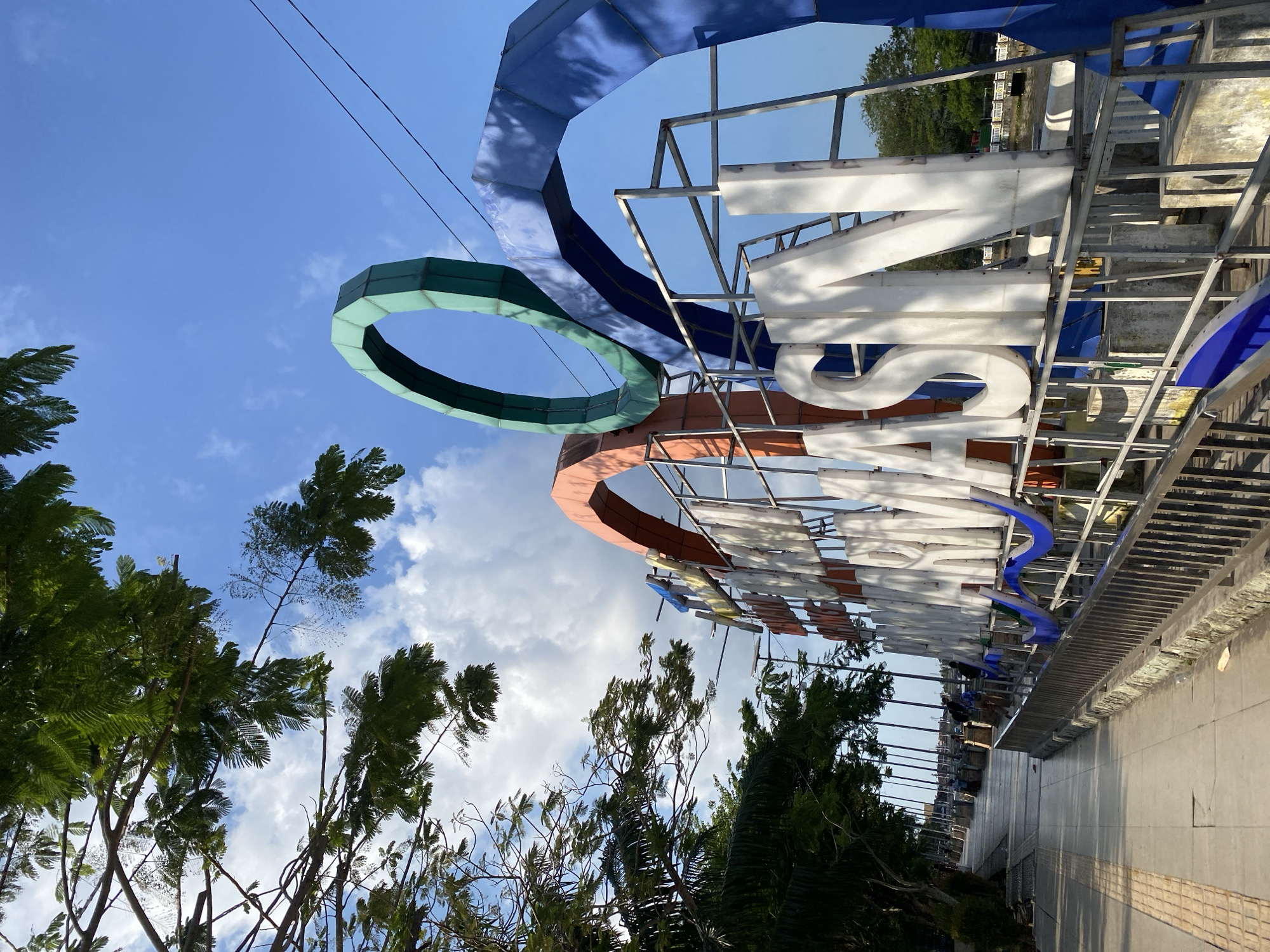 The image shows a large, white, three-dimensional sign that reads "MARTAPURA" against a bright blue sky with scattered clouds. The sign is framed by abstract, colorful arches in orange, green, and blue, creating a modern and artistic visual. Lush green trees with feathery leaves overhang the scene from the left, their branches casting shadows on the sign and the sky.
In the foreground, a paved walkway runs along the left side of the image, bordered by a metal railing and a textured stone wall. The walkway appears to be a public area, possibly a promenade or a park, with a few indistinct figures of people visible in the distance further along the path. The railing in front of the sign has a blue wavy decorative element attached to it.
The sign and its architectural framing are positioned next to a body of water, indicated by the reflection on the water's surface and the presence of a bridge in the background. The overall scene depicts an outdoor public space in Banjarmasin, Indonesia, likely a waterfront area, with the prominent sign serving as a landmark. The lighting suggests it is daytime, with the sun casting strong shadows.
