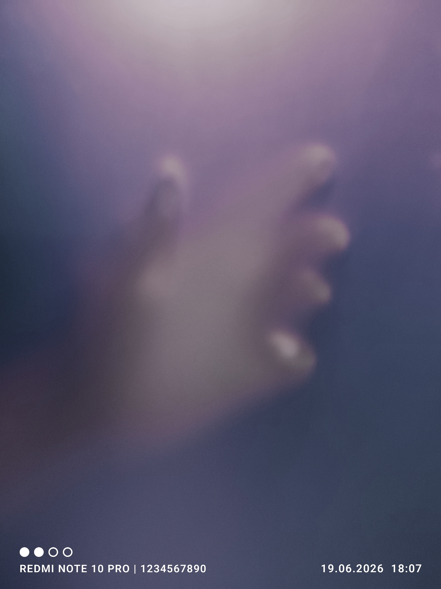 This media file is a photo of a person's hand seen through a blurred, translucent surface. The background is a gradient of purple and dark grey, with a soft, diffused light creating an ethereal effect. The hand appears to be reaching out or resting against the surface, with the fingers partially visible. The overall mood is somewhat mysterious and abstract due to the heavy blurring.
Visible text in the image includes "REDMI NOTE 10 PRO | 1234567890" at the bottom left, and "19.06.2026 18:07" at the bottom right, indicating the device used and the date and time the photo was taken. There are also four white dots at the bottom left, commonly used in phone interfaces to indicate progress or navigation. The time of day is indicated by the timestamp as 18:07, which is early evening. No specific weather or location cues are discernible beyond the indoor or studio-like lighting. The emotion conveyed is ambiguous due to the abstract nature of the image.